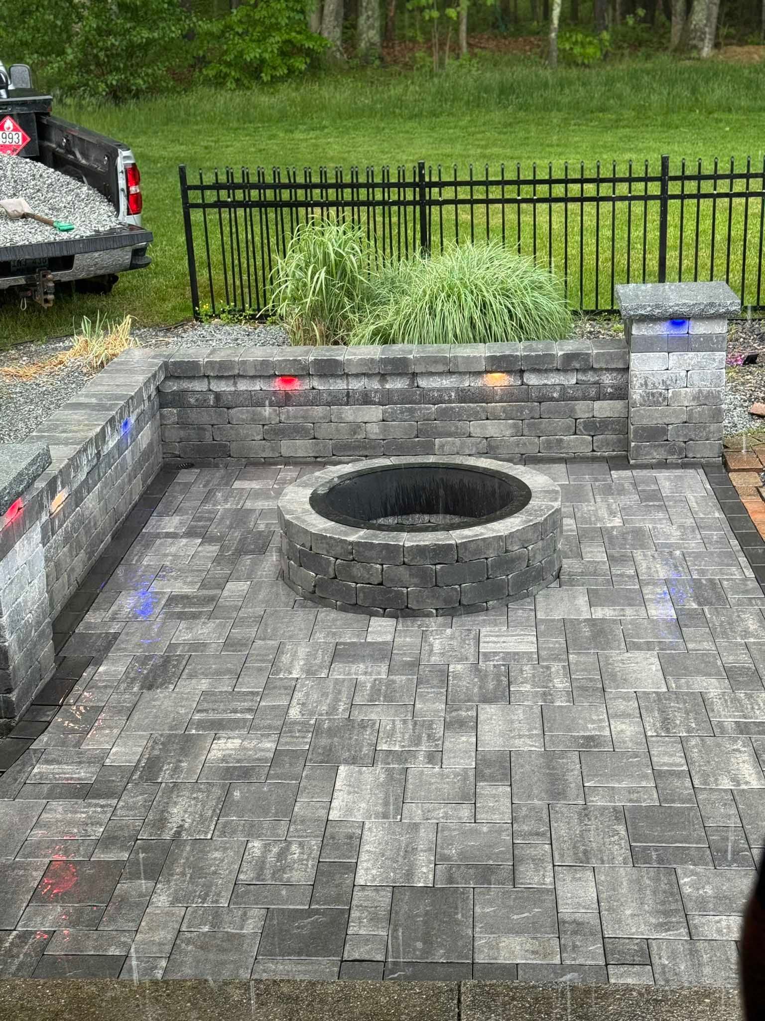 A circular stone fire pit sits on a gray paver patio, surrounded by a matching retaining wall and a black metal fence.