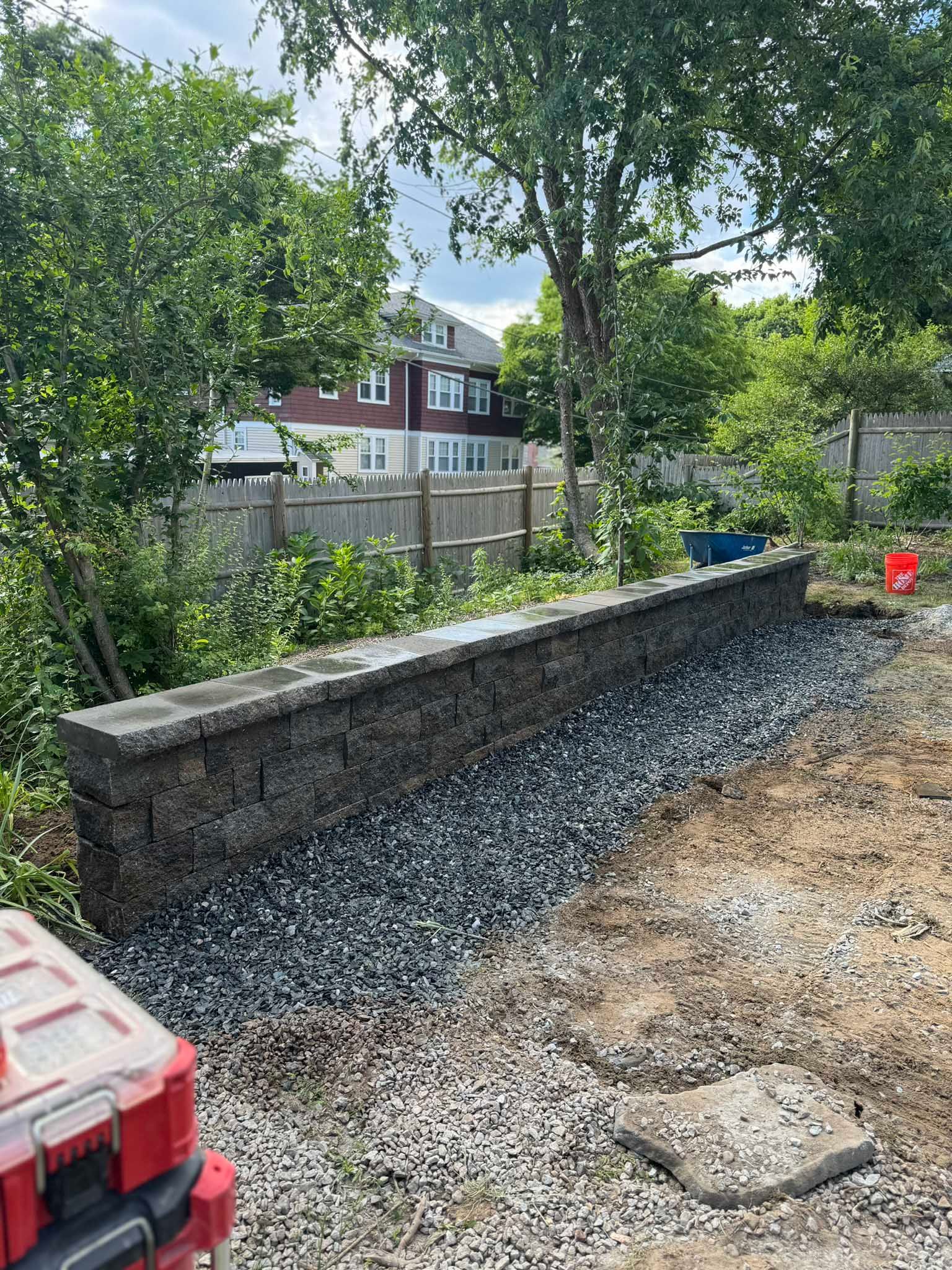 A newly constructed stone retaining wall sits in a backyard filled with gravel, bordered by a wooden fence and trees.
