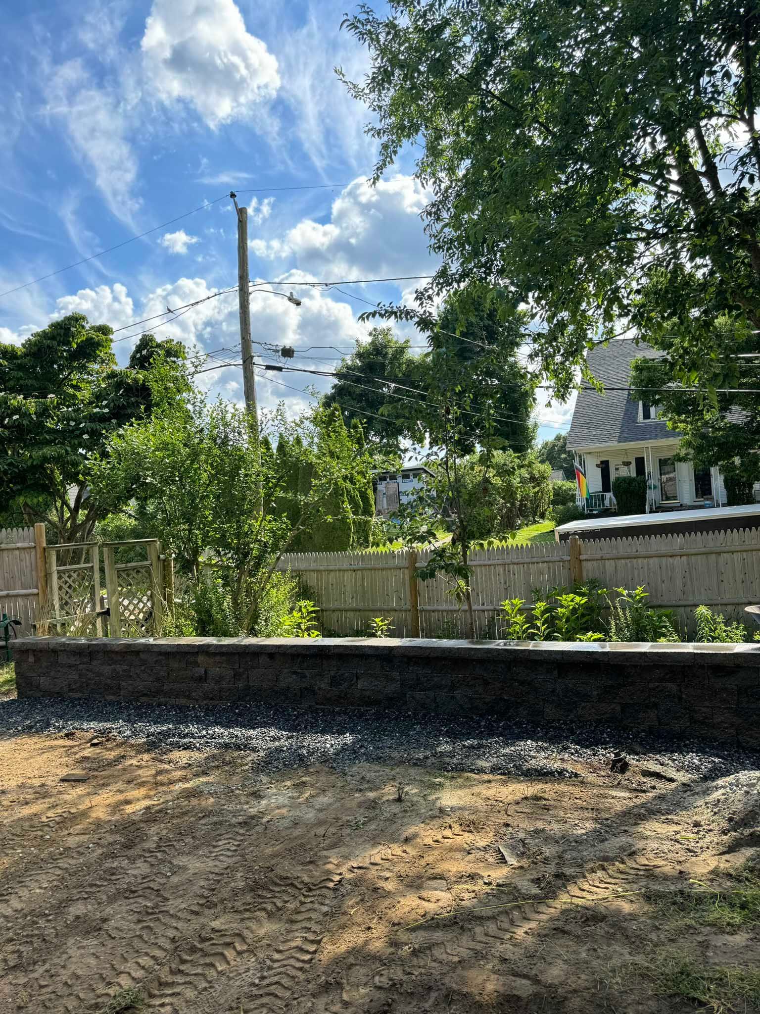 A stone retaining wall sits in front of a wooden fence and lush trees under a partly cloudy blue sky.