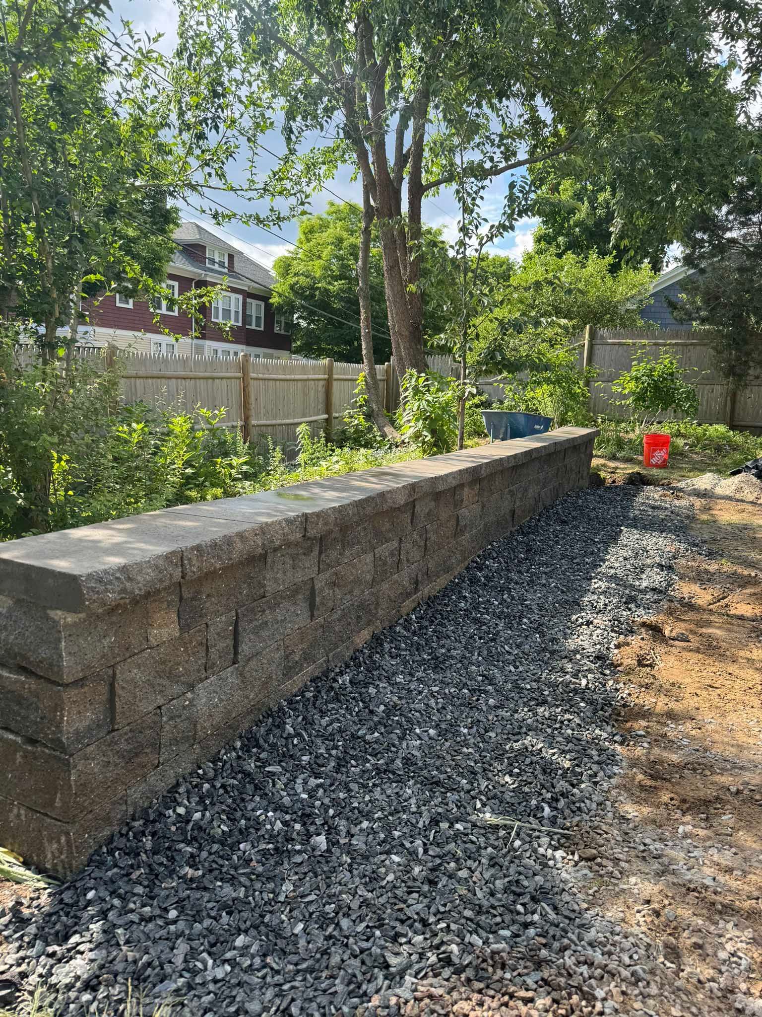 A newly built, textured concrete retaining wall stands next to a strip of gravel in a sunny backyard with trees and a fence.