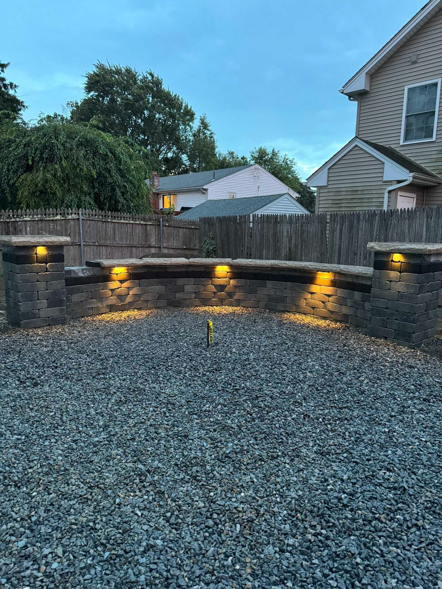 A curved stone retaining wall with integrated warm lighting, set against a wooden fence and gravel backyard at dusk.