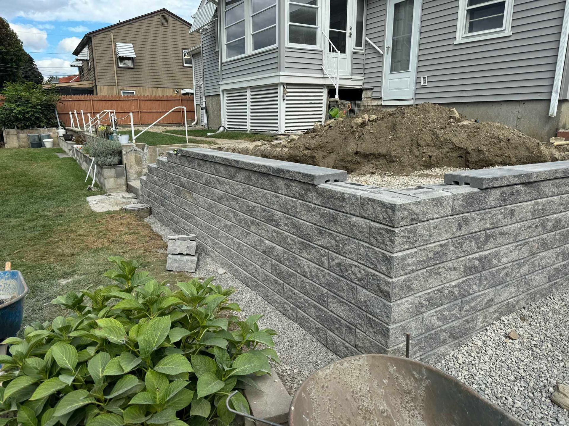 A newly built gray concrete retaining wall sits in front of a residential house with a pile of dirt behind it.