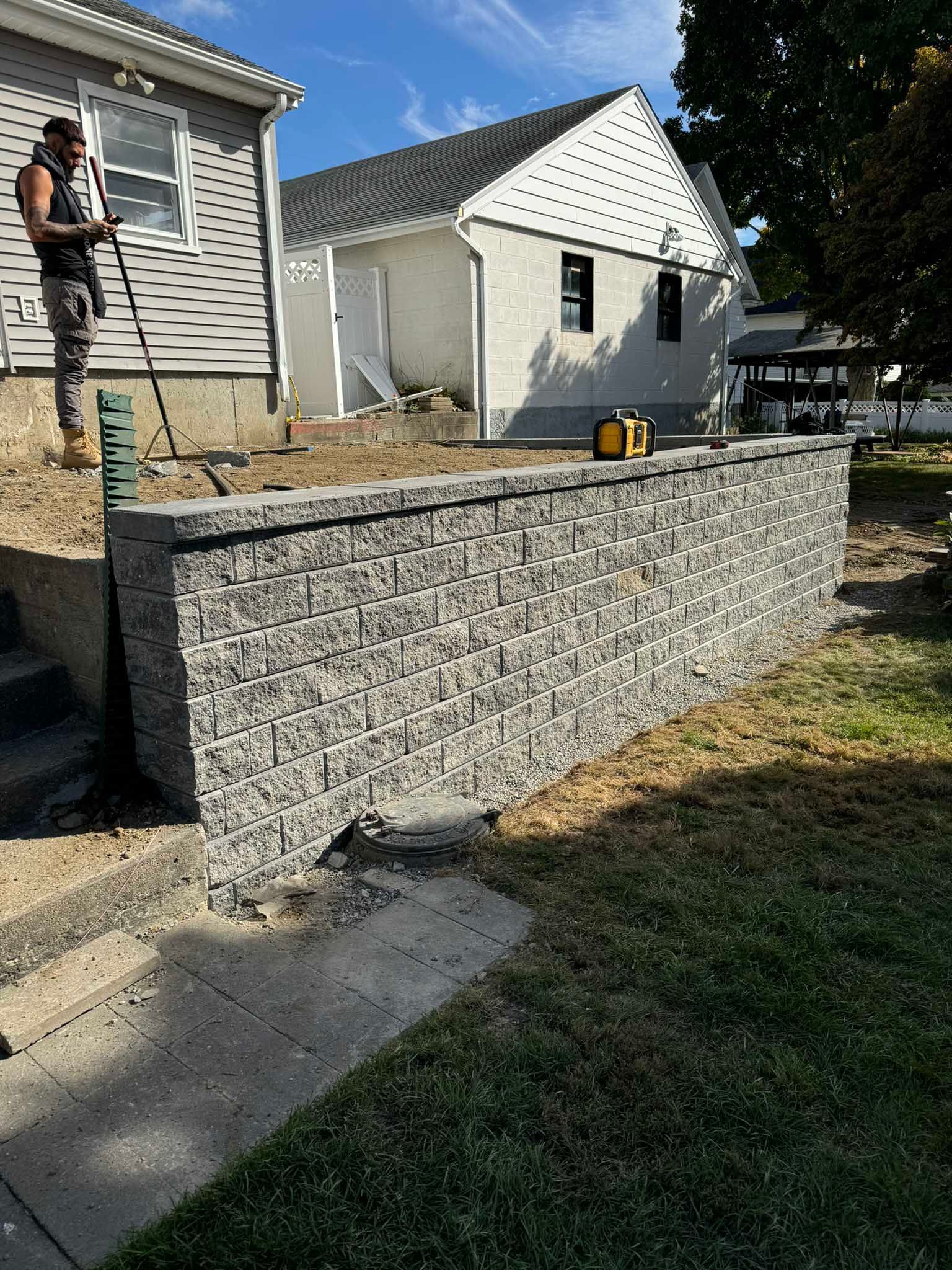 A worker stands near a newly built gray stone retaining wall in front of two residential houses on a sunny day.