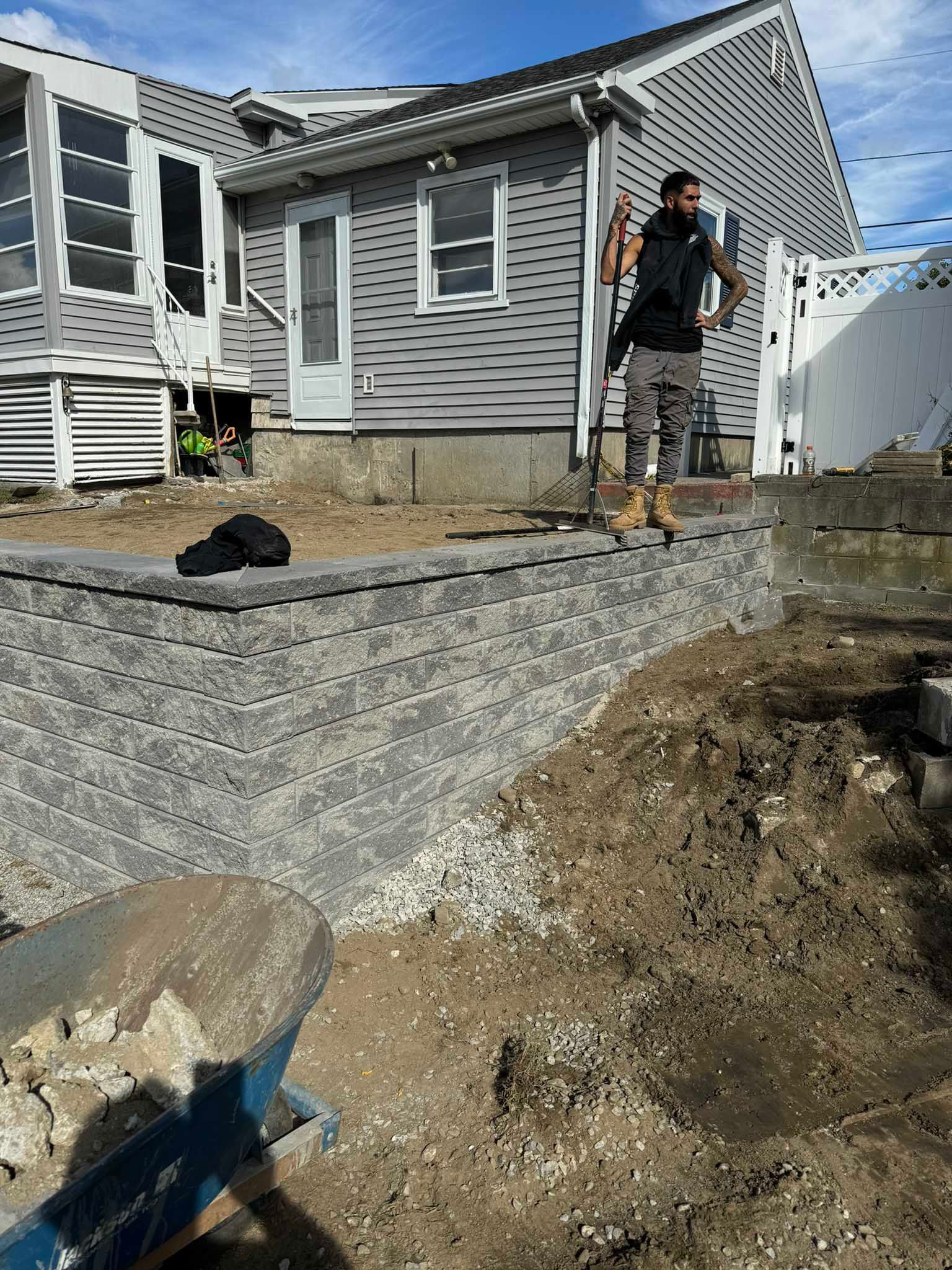 A construction worker stands on a newly built gray stone retaining wall in a yard beside a house with gray siding.