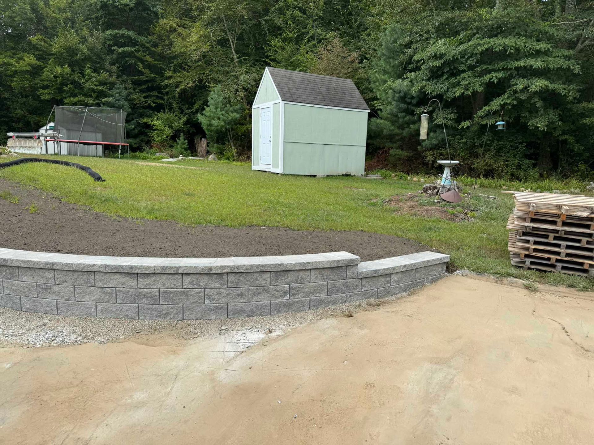 A low, gray stone retaining wall stands in a yard, bordered by a dirt patch, a small shed, and a stack of wooden pallets.