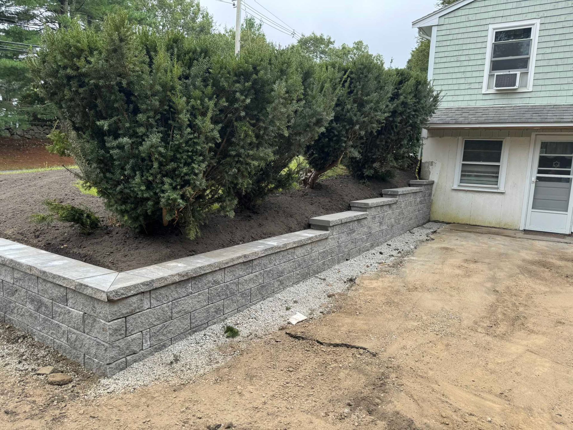 A stone retaining wall sits in front of a row of large, dense green bushes next to a light-green siding house.