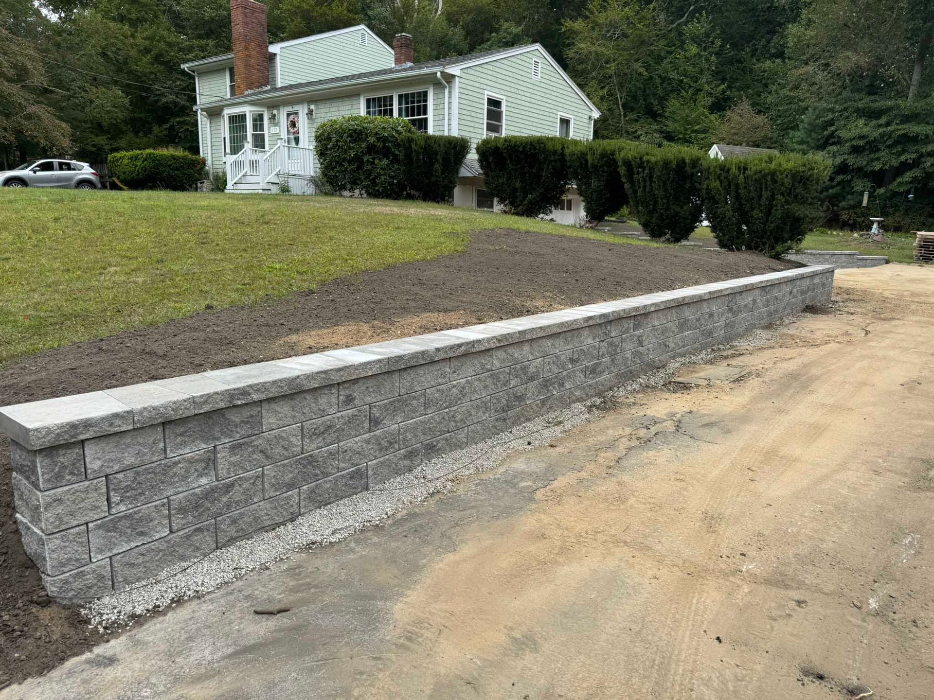 A gray, segmented stone retaining wall runs alongside a dirt driveway in front of a house with a green lawn.