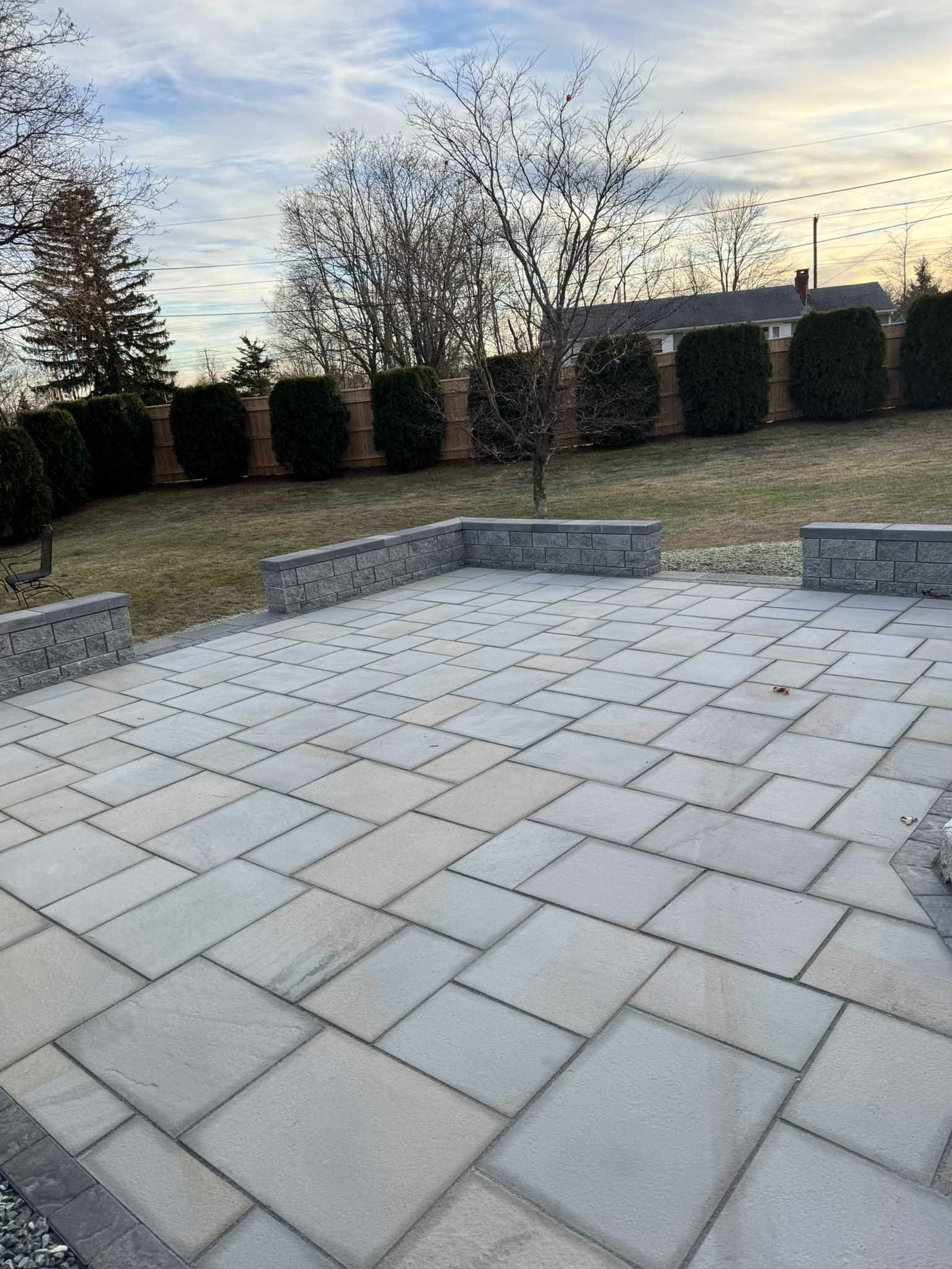 A stone paver patio with low retaining walls in a backyard with a line of hedges and a tree in the background.