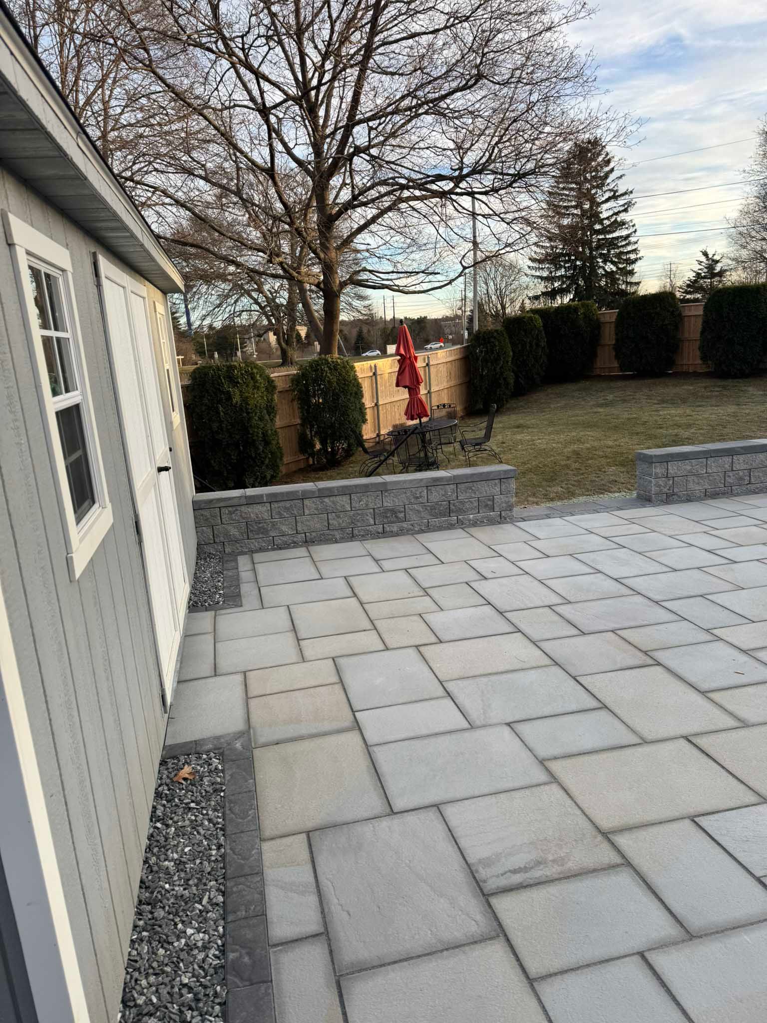 A grey shed beside a large, light-grey stone patio with a retaining wall, overlooking a backyard lawn with trees.