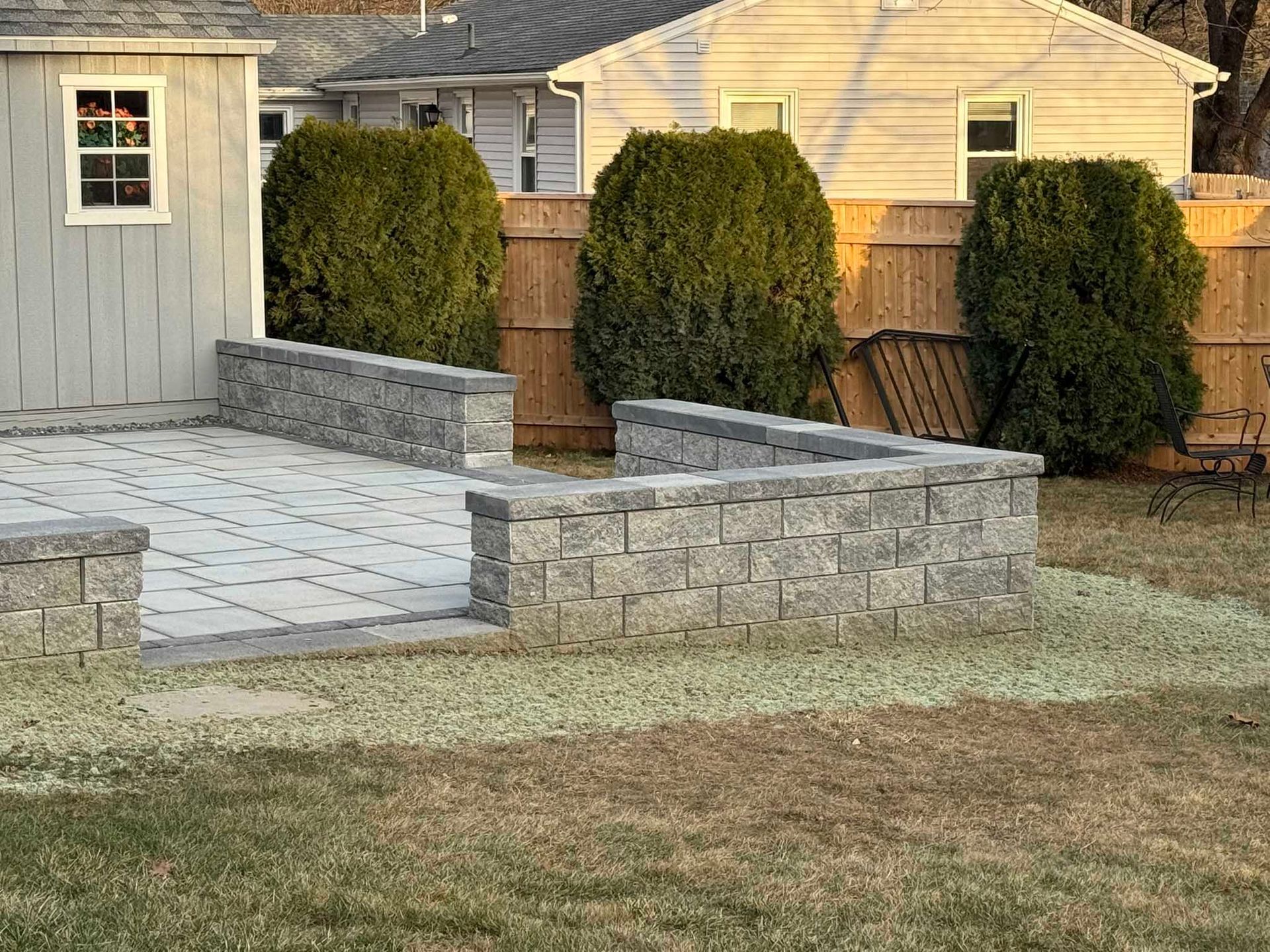 A grey stone retaining wall surrounds a light-colored paved patio next to a shed and a wood fence in a backyard.