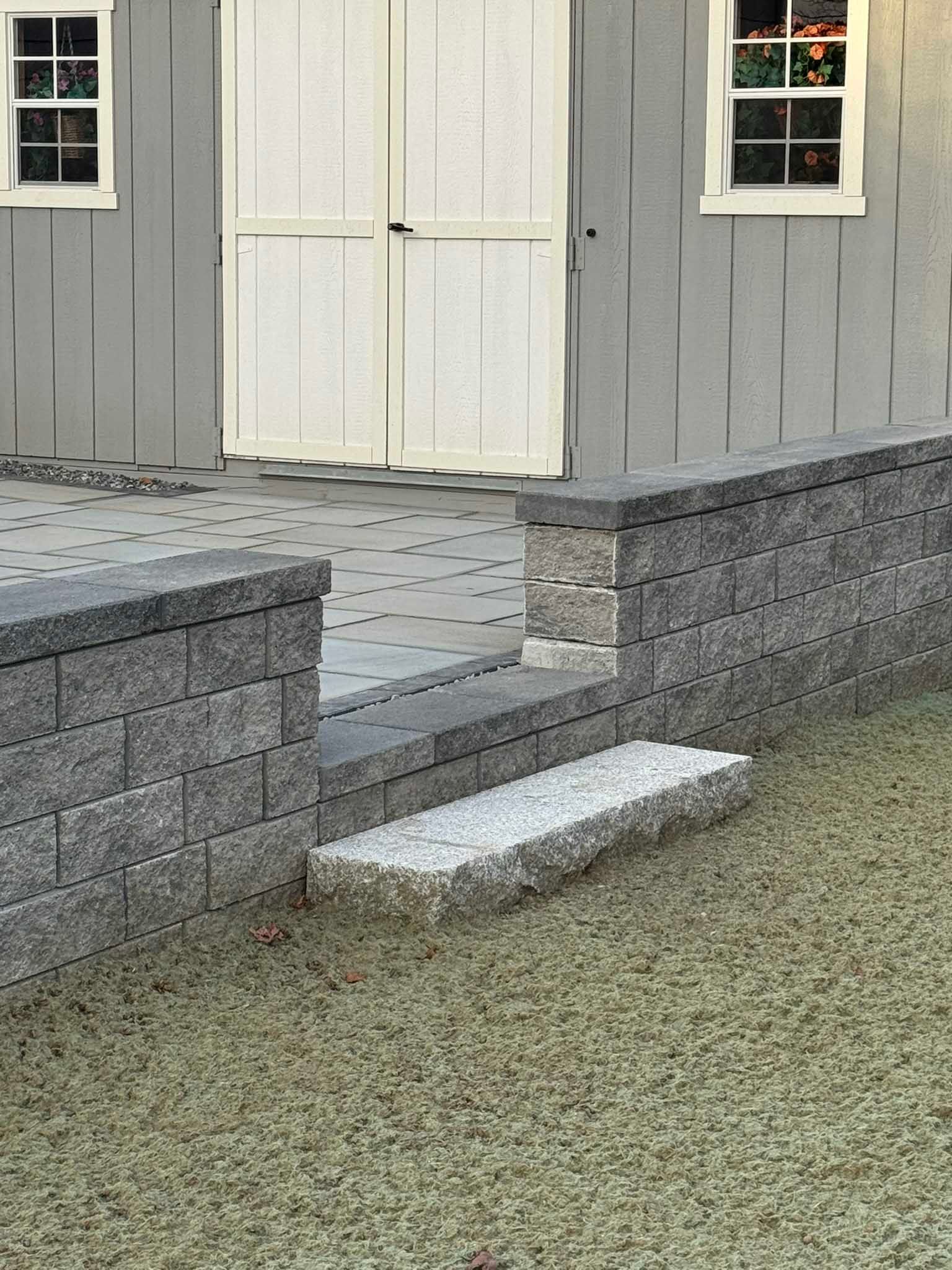 A gray shed with white doors sits above a stone retaining wall featuring a single wide step leading up to a paved patio.