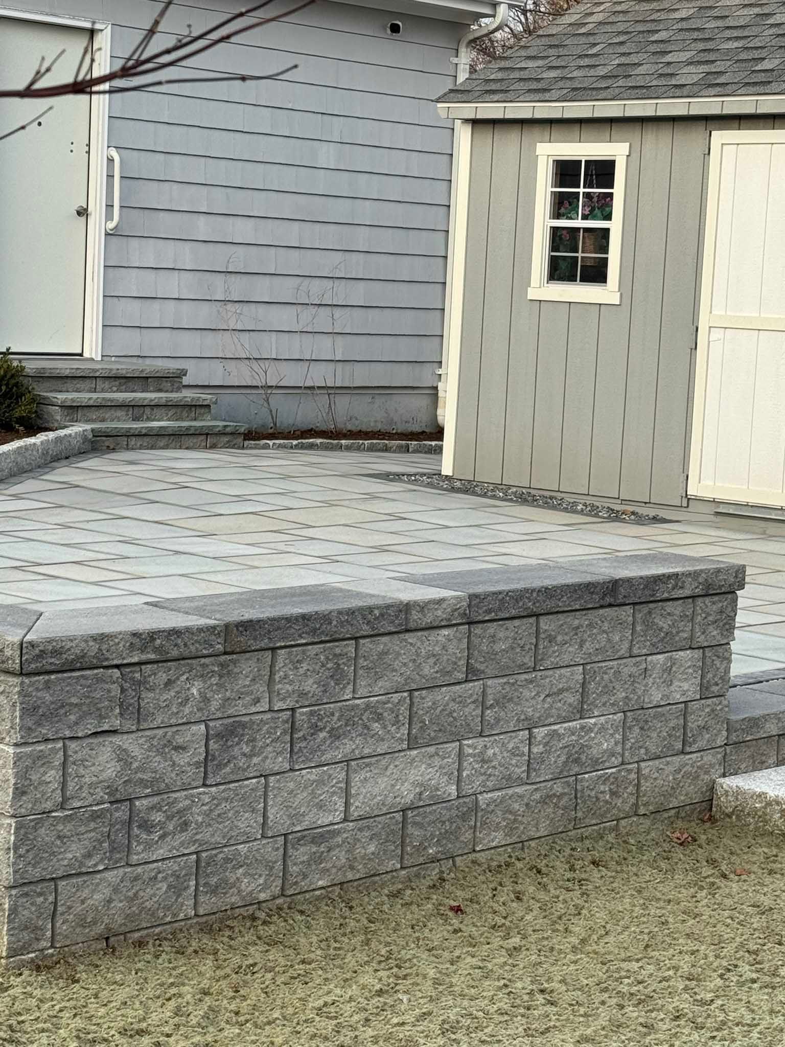 A gray stone retaining wall stands in front of a paved patio, a small storage shed, and a house entrance.