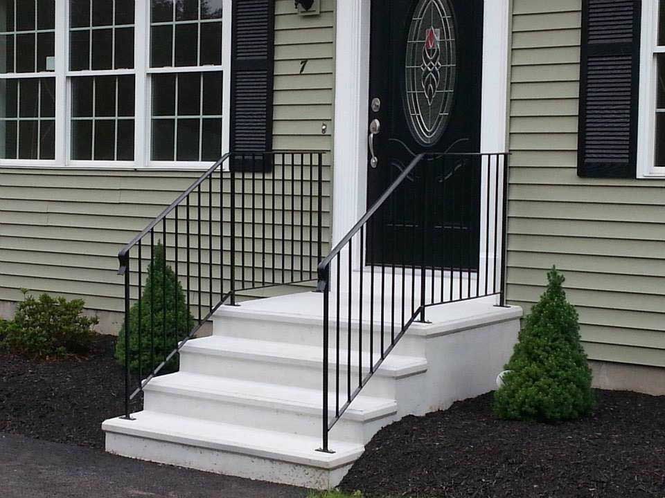 A black metal handrail on white concrete steps leading to the front door of a home with green siding.