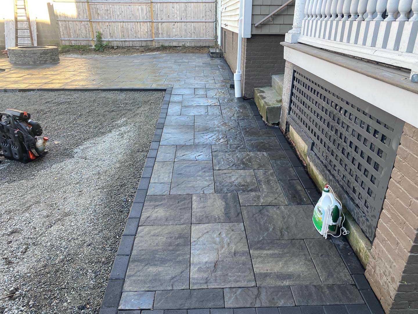 A stone patio path runs alongside a house deck, bordering a gravel area with a leaf blower in the background.