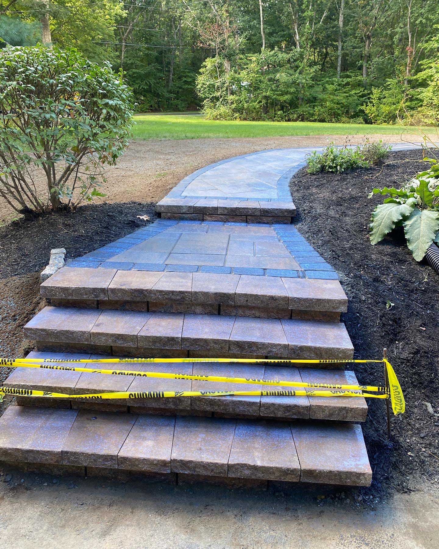 A set of four stone steps leading to a curved paved walkway, blocked by yellow caution tape, surrounded by mulch.