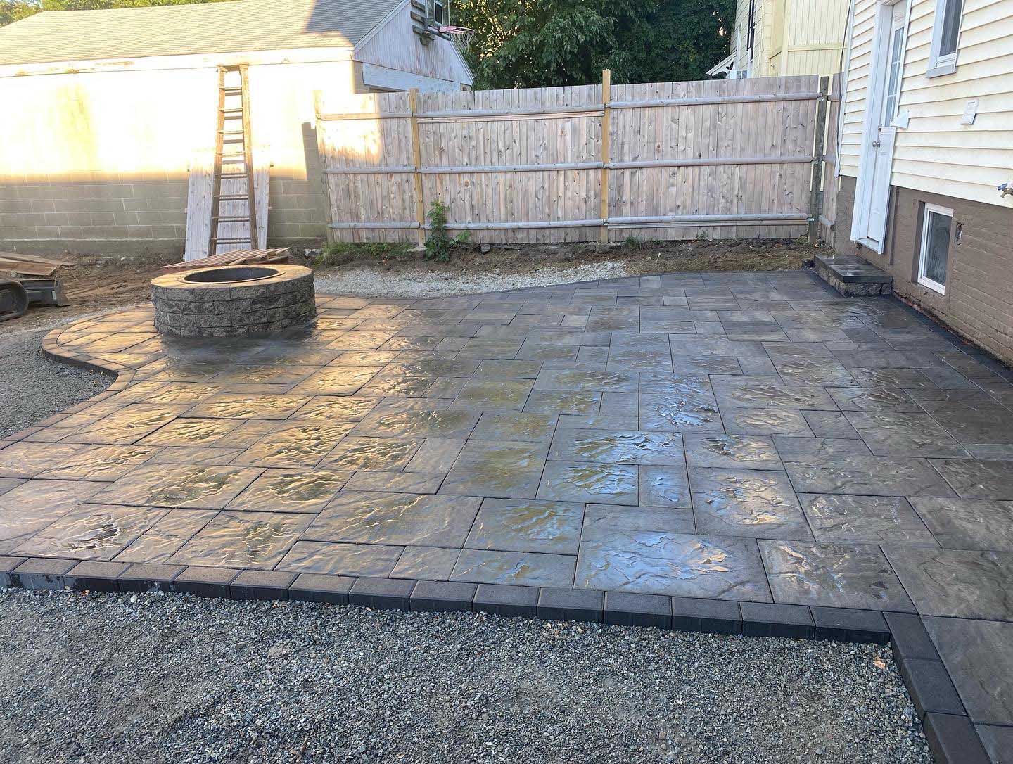 A backyard patio made of textured, grey rectangular pavers featuring a circular stone fire pit, set beside a house.