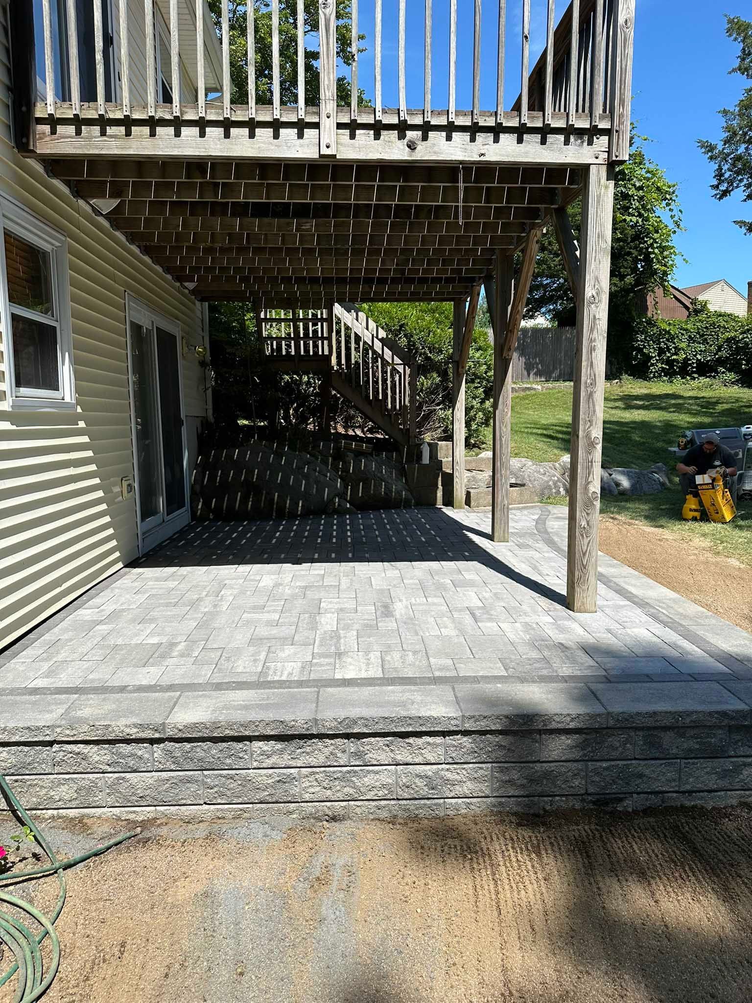 A newly installed gray paver patio with a stone retaining wall sits underneath a wooden deck attached to a house.
