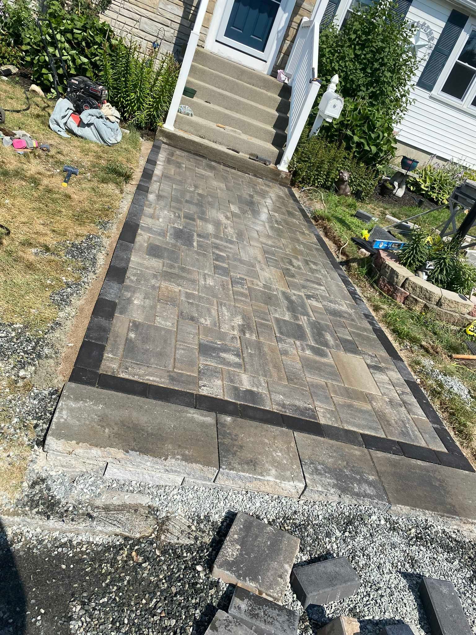 A newly installed stone paver walkway with a dark border leads up to a set of concrete steps in front of a house.