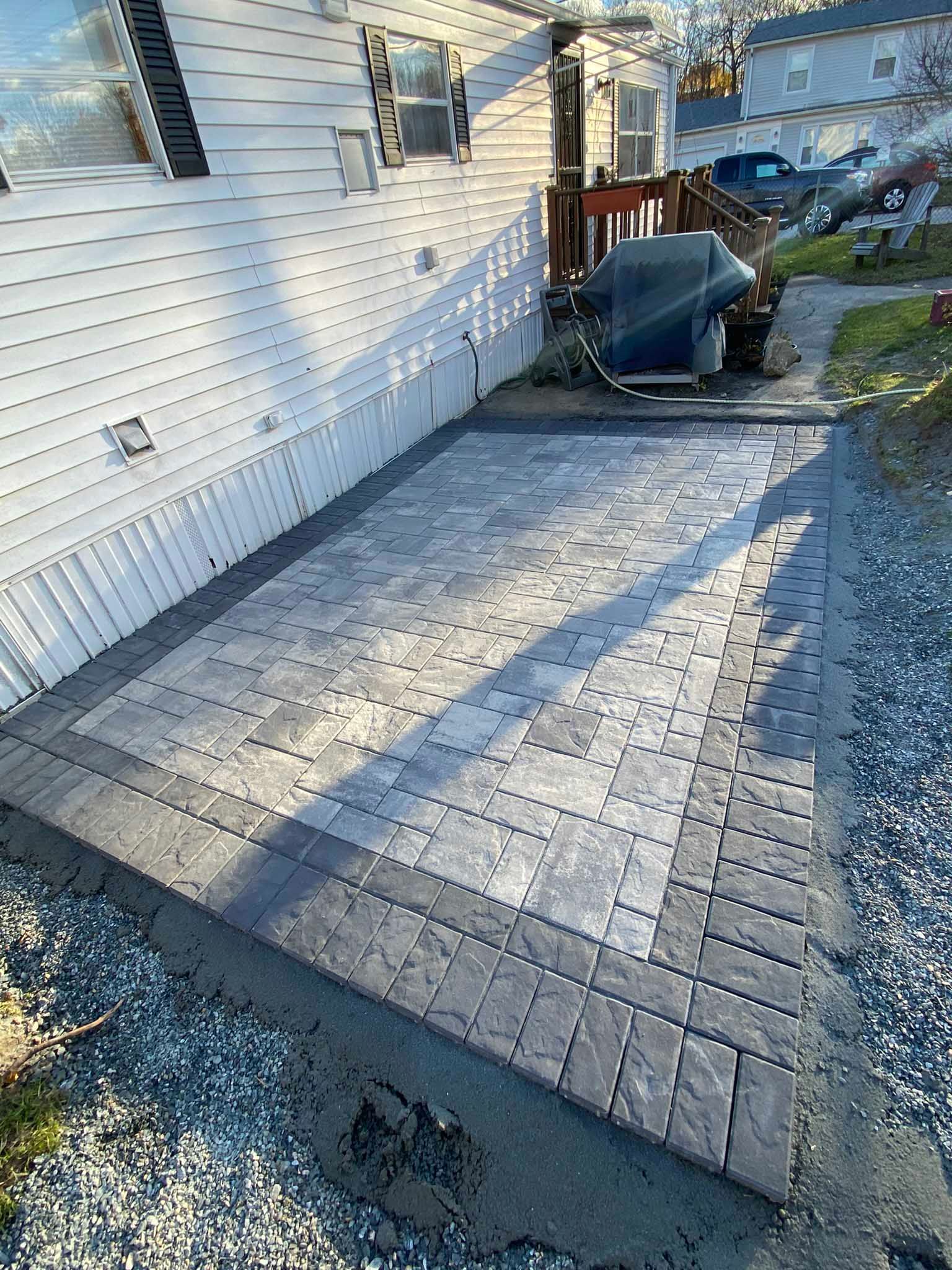 A newly installed gray paver patio with a darker border sits beside a white-sided mobile home.