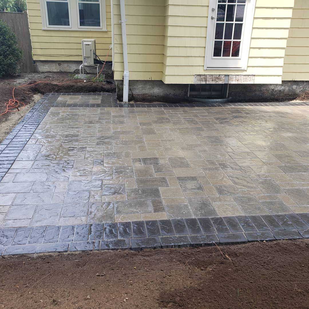 A stone patio with a light gray center and dark gray border sits beside a house with yellow siding and a white door.