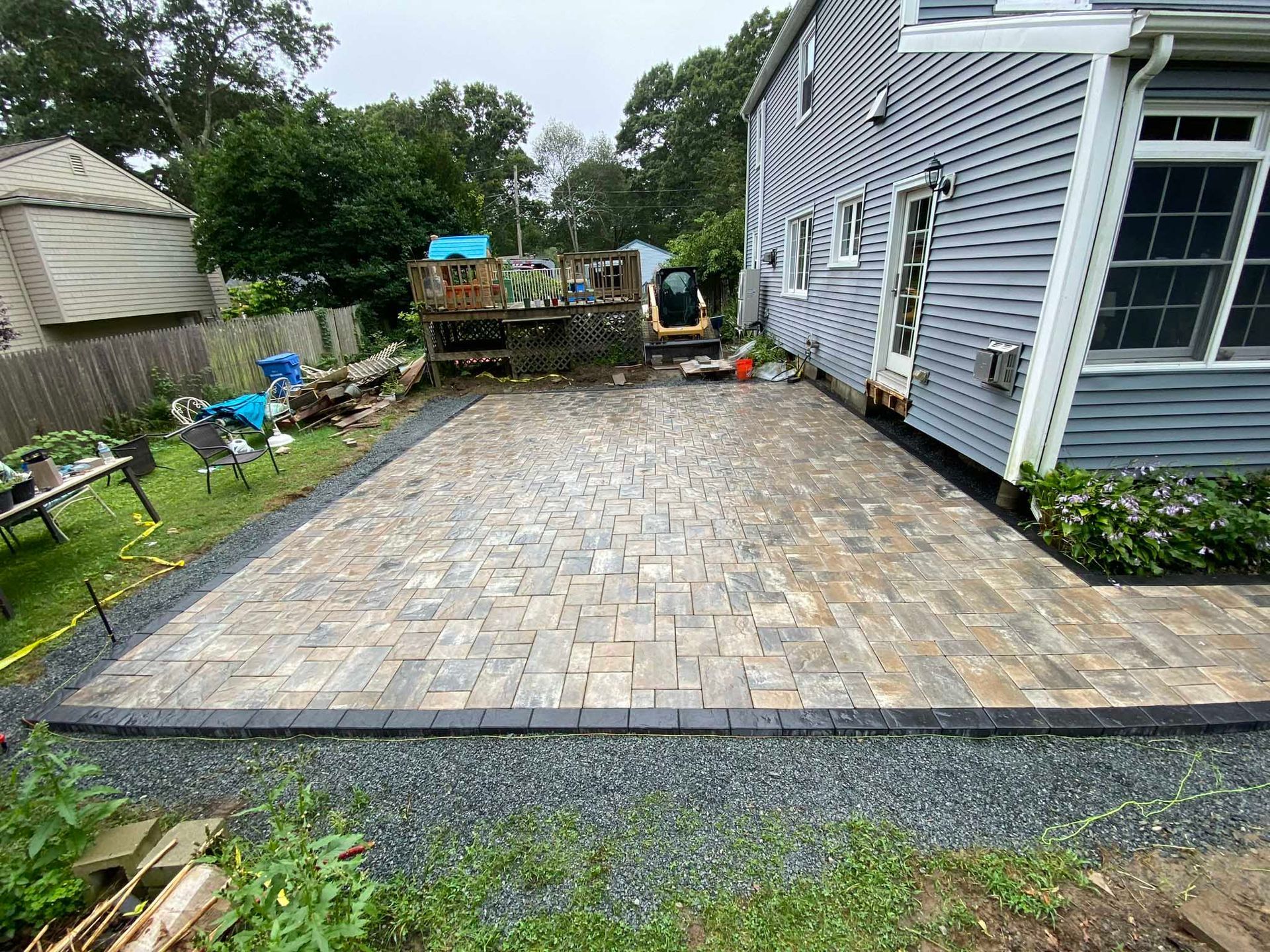 A newly installed stone paver patio sits adjacent to a grey house, bordered by dark gravel and a grassy yard.