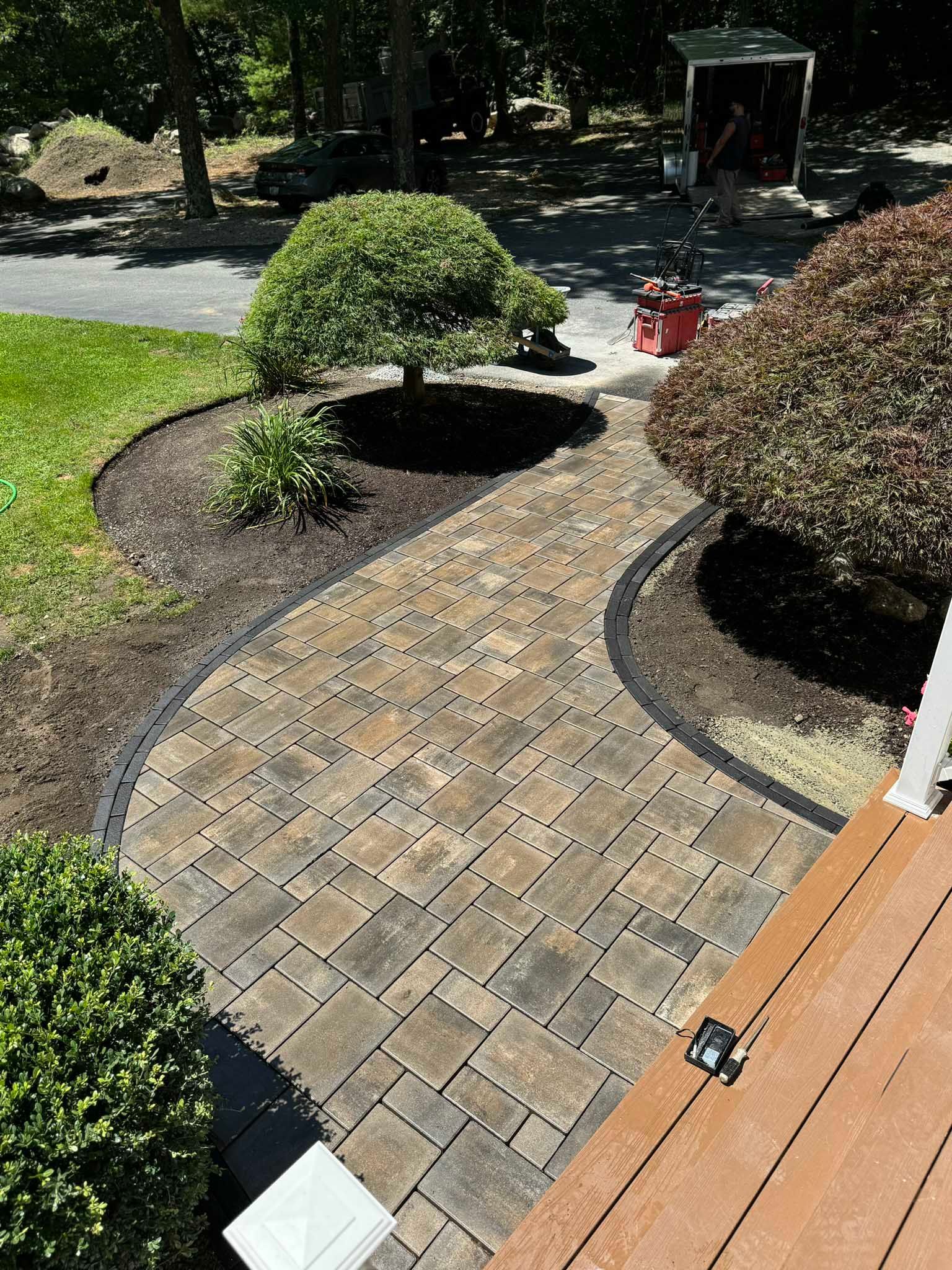A curved paver walkway leads from a wooden deck past landscaping, trees, and a small patch of grass toward a driveway.