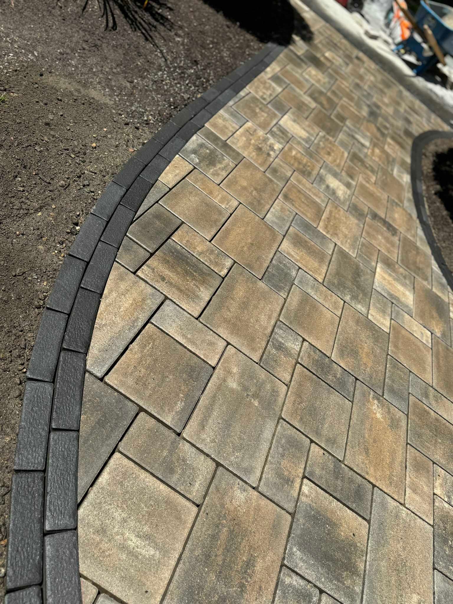 A paved walkway with rectangular tan and grey pavers, bordered by a curved edge of dark charcoal bricks.