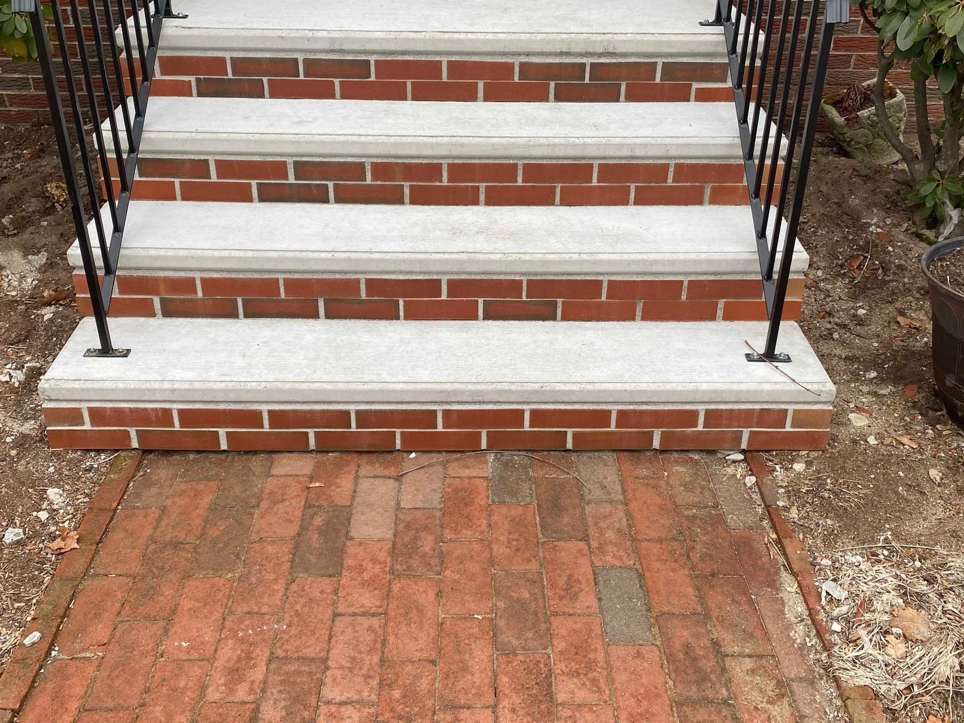 A brick walkway leads to three stone-topped brick steps with black metal railings in an outdoor setting.