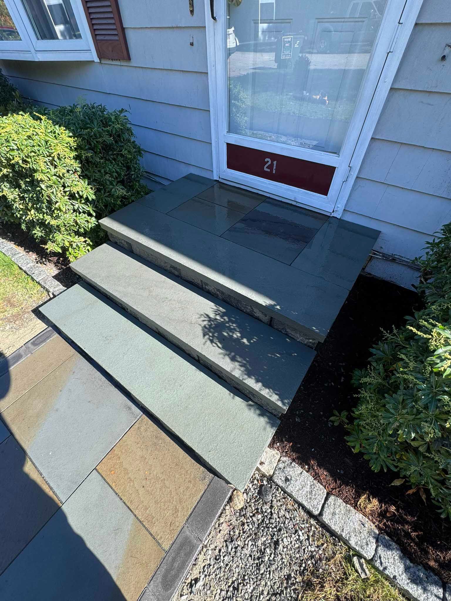 A set of three dark stone steps leading up to a front door with a white frame and a dark red panel with the number 21.