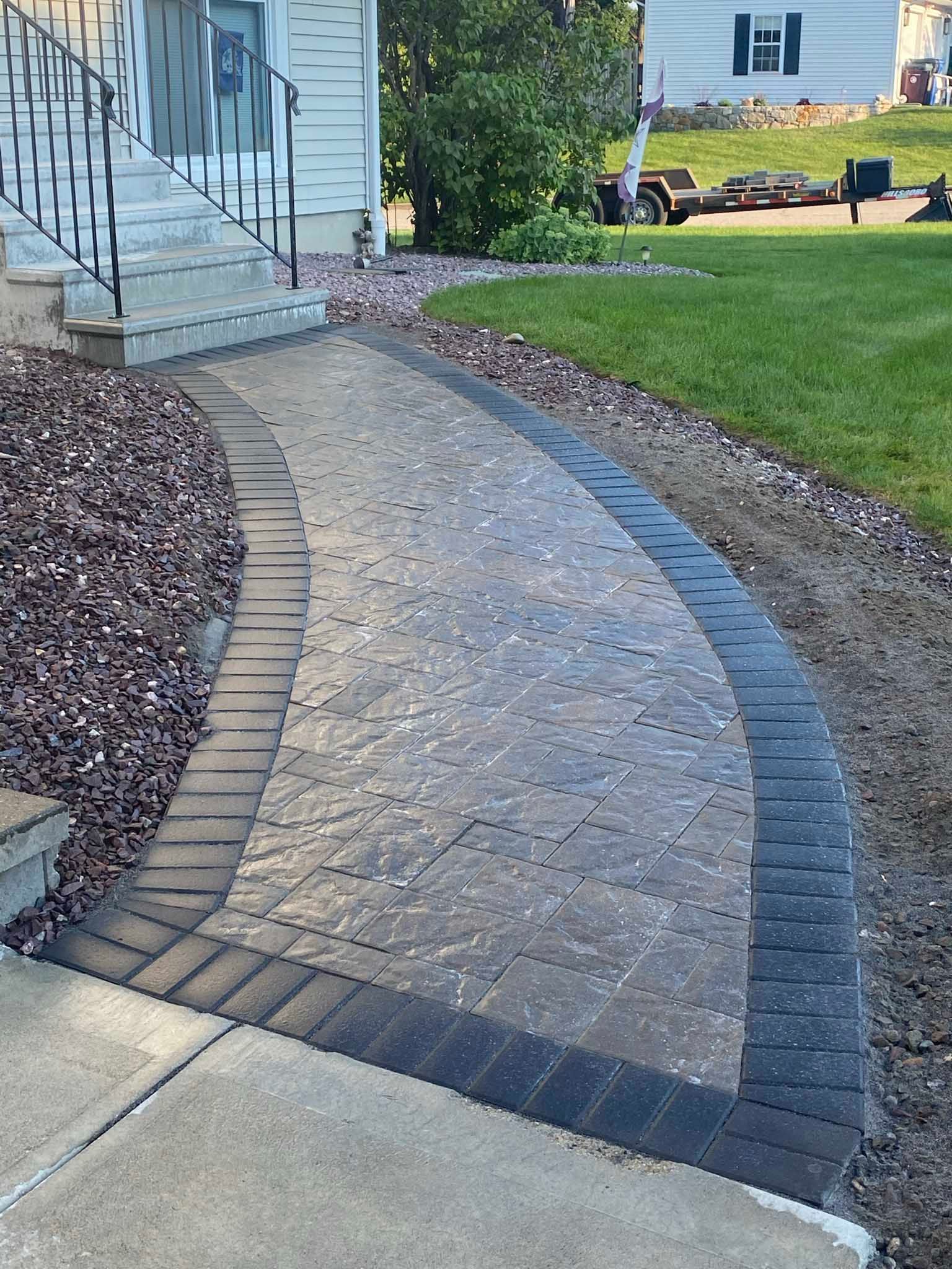 A paved walkway with a tan textured center and dark gray border leads from a concrete sidewalk to white house steps.