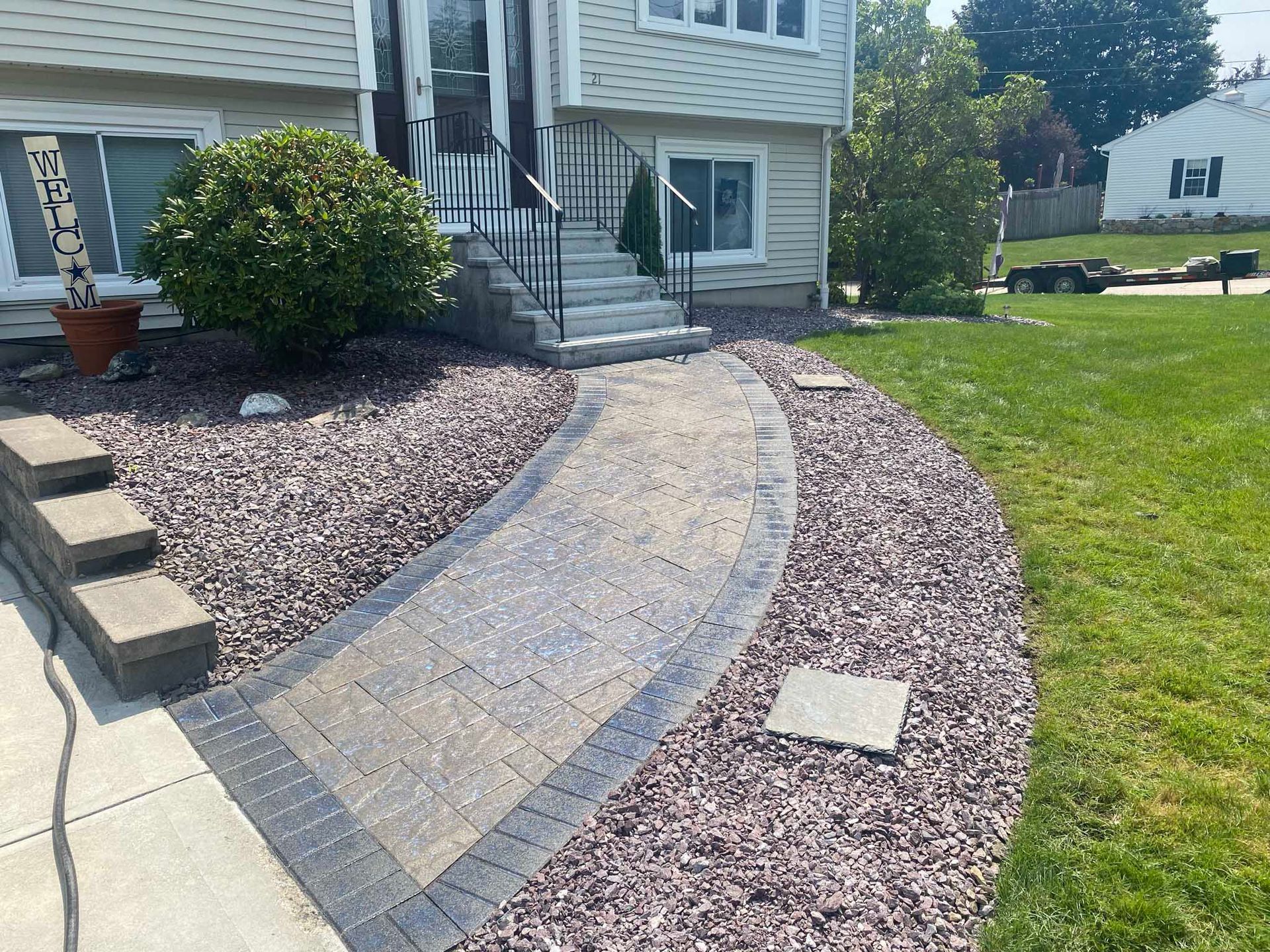 A curved paver walkway leads to a house entrance with gray stone steps, surrounded by purple stone mulch and greenery.