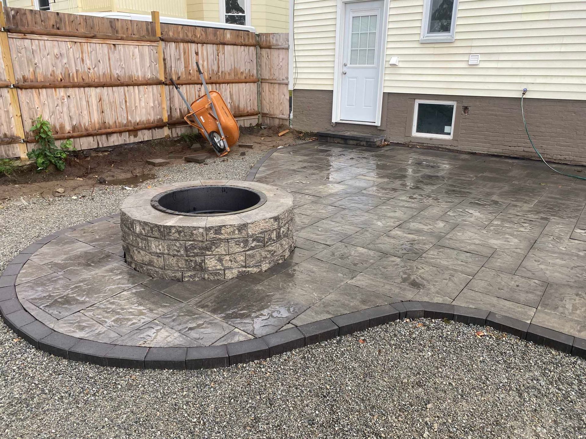 A backyard patio featuring a circular stone fire pit, surrounded by gray pavers and edged with darker bricks on gravel.