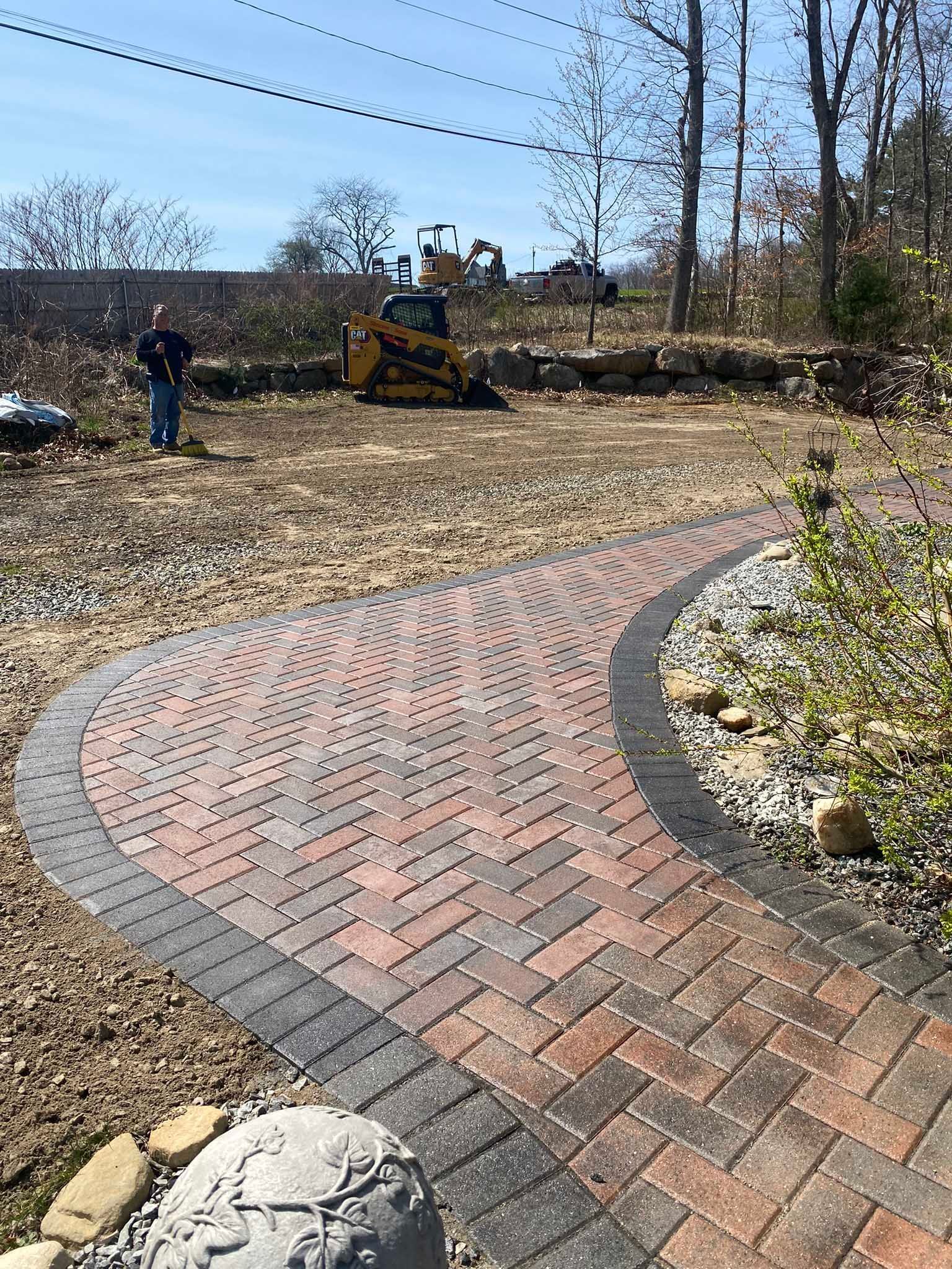 A person stands near a curved brick walkway with a decorative pattern, leading toward construction work in a yard.