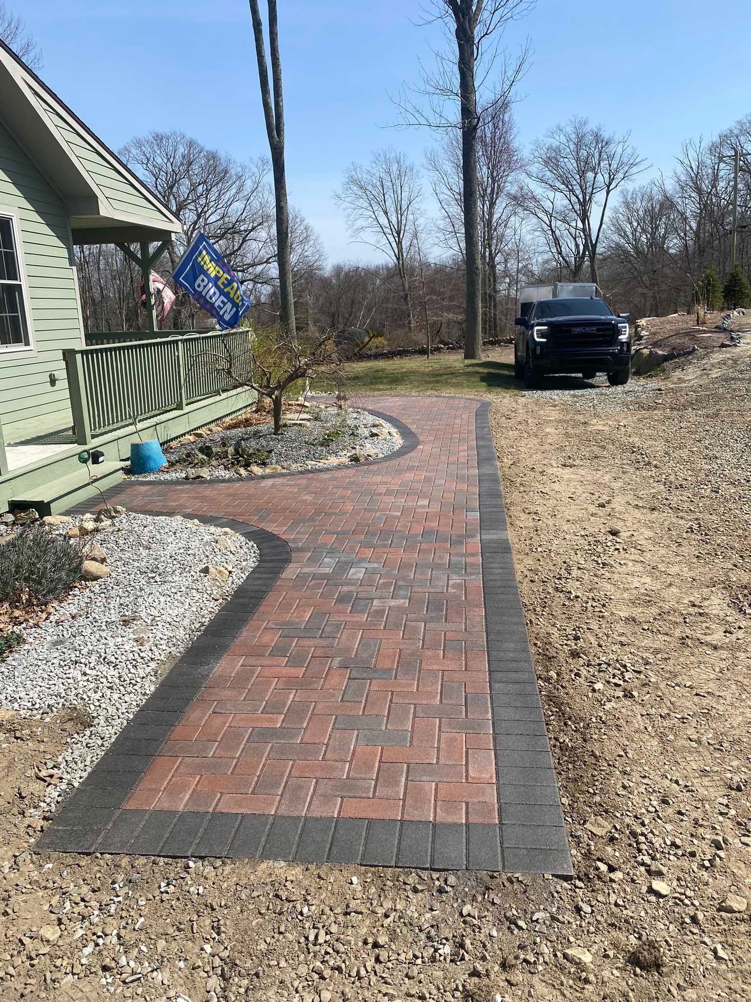 A walkway made of red brick pavers with a dark gray border leads to the porch of a light green house on a sunny day.