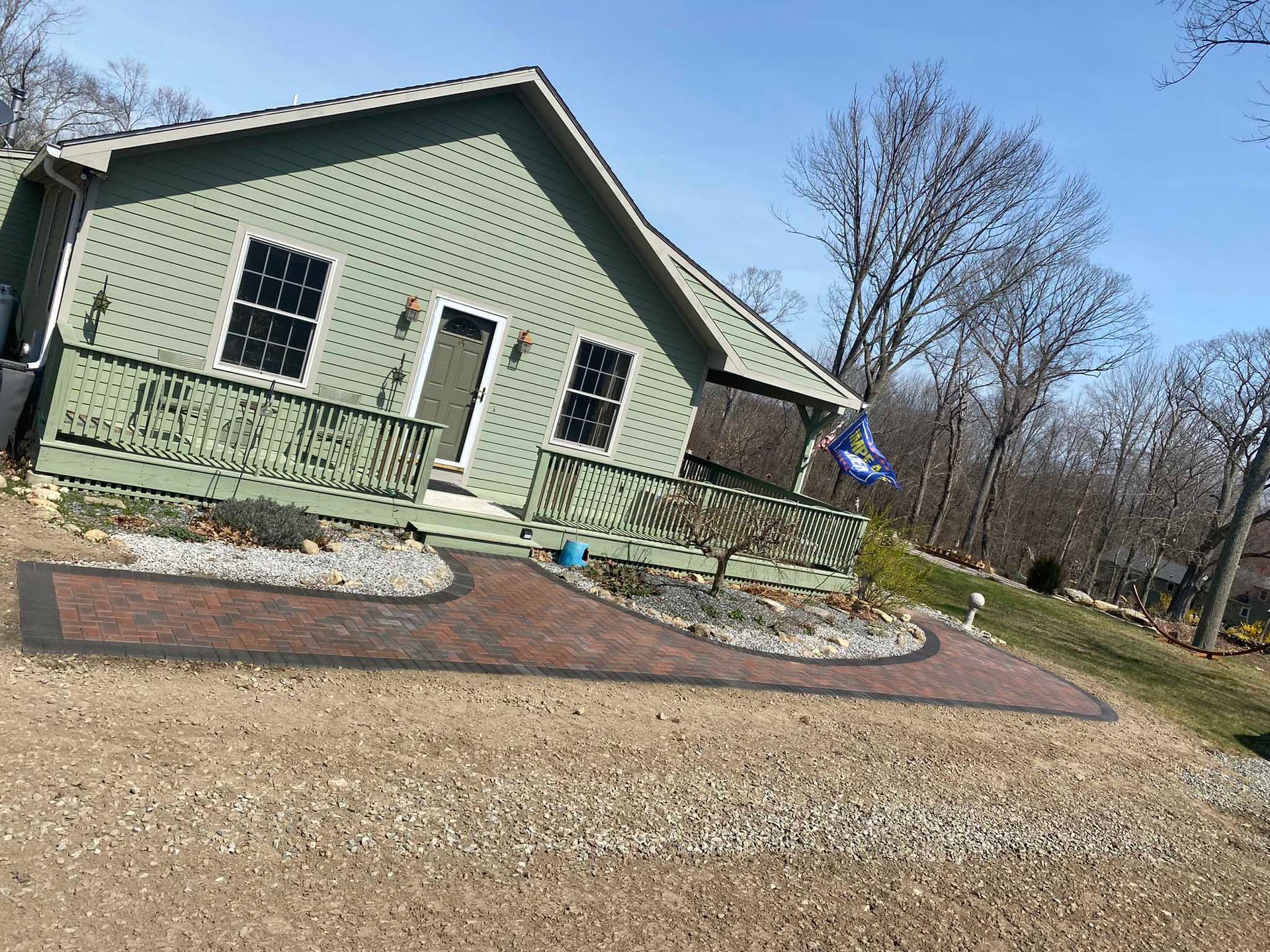 A sage green cottage with a porch and a paver walkway, set in a sunny, wooded outdoor area.