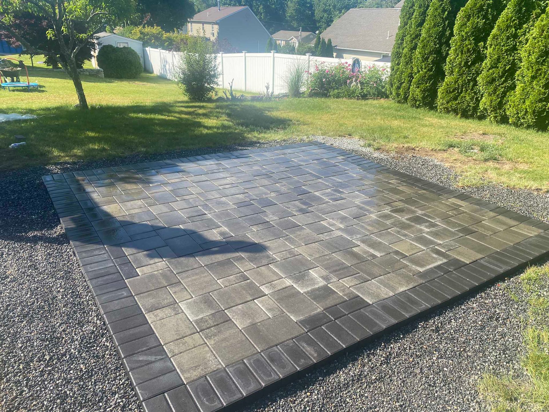 A rectangular backyard patio made of grey and black pavers, surrounded by a gravel border on a sunny day.