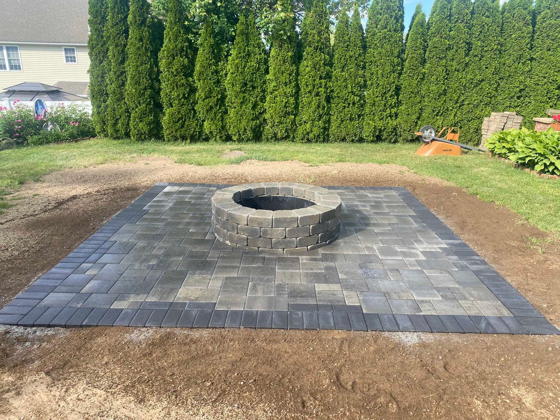 A stone fire pit sits in the center of a rectangular gray paver patio set in a backyard lawn with a tall evergreen hedge.
