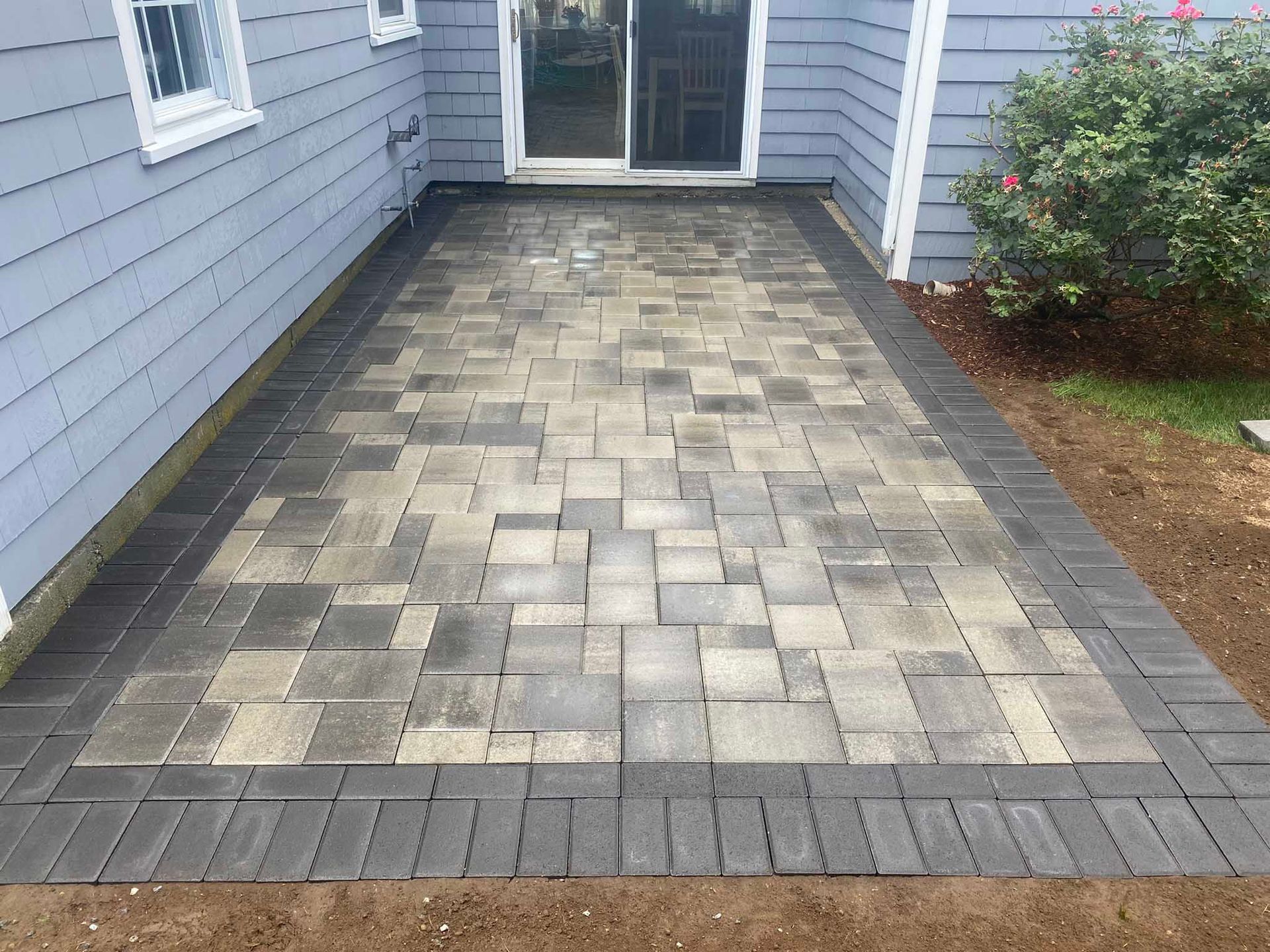 A rectangular patio made of rectangular gray and tan pavers with a dark charcoal border, situated beside a blue house.