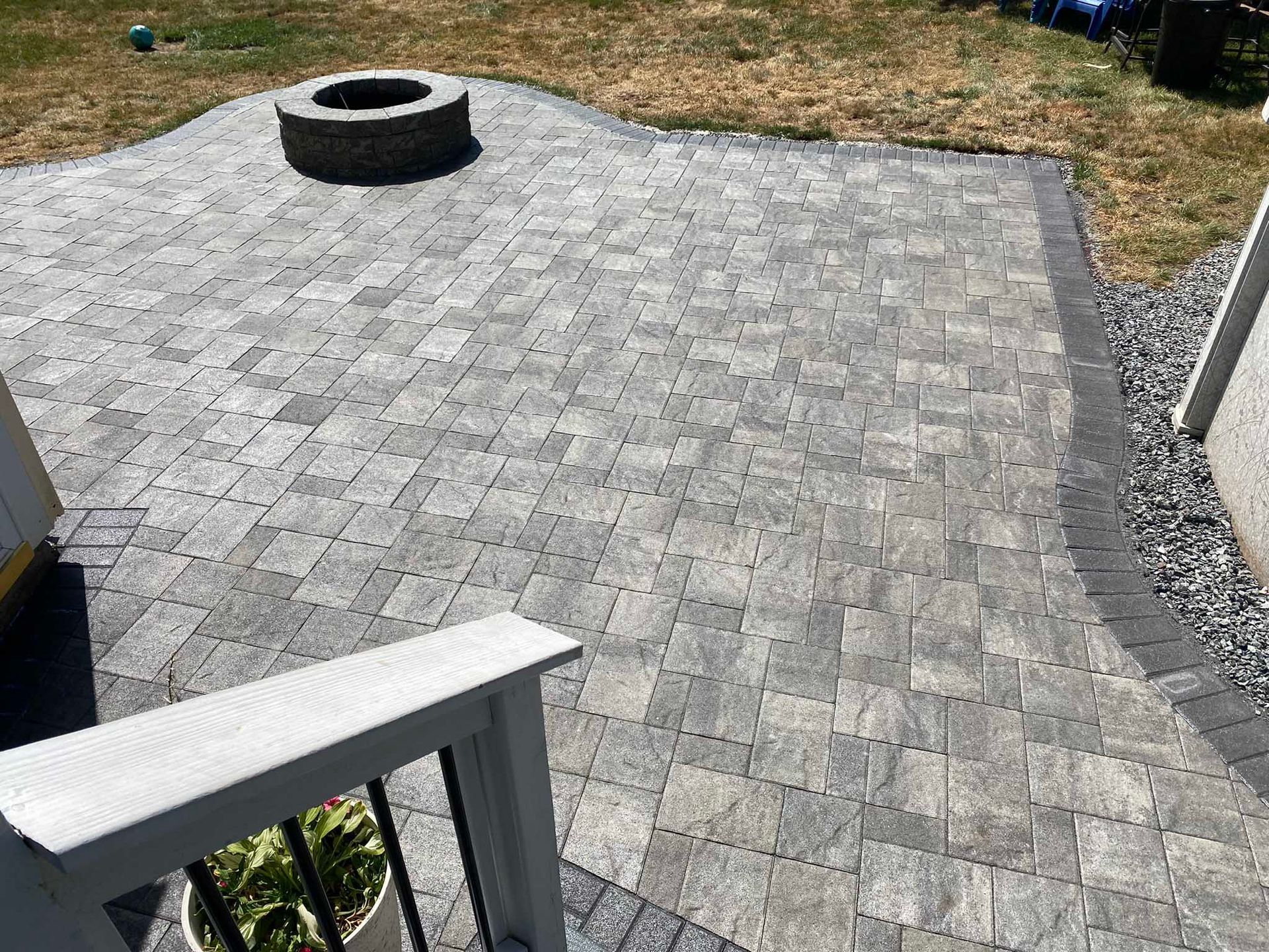 A patio paved with gray and charcoal rectangular stones, featuring a matching circular fire pit in the grass-lined yard.