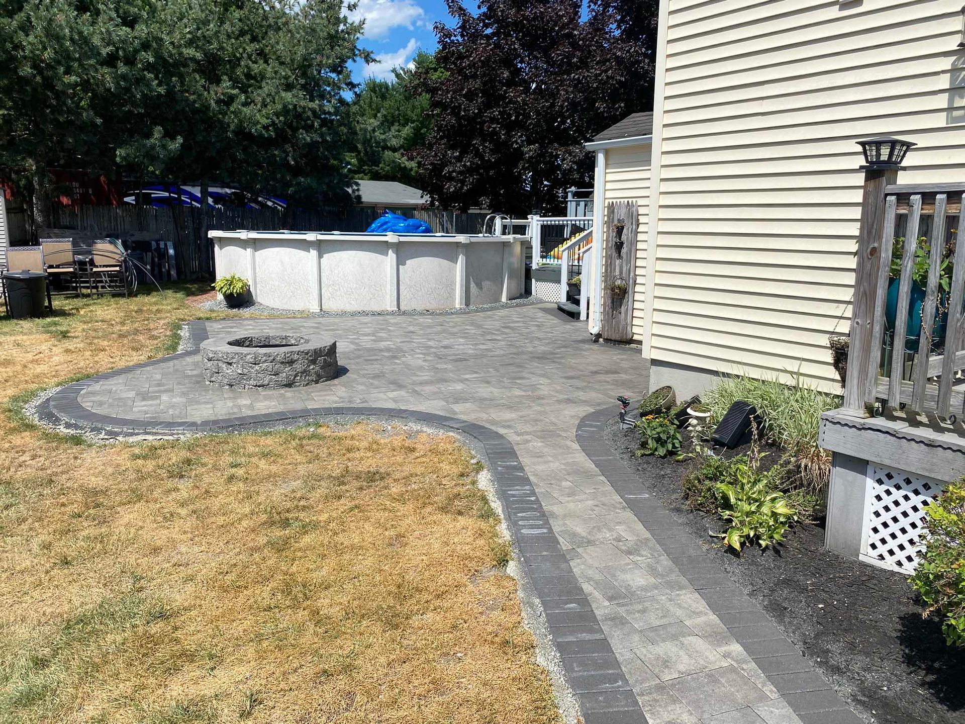 A paved stone patio with a fire pit, adjacent to an above-ground pool, next to a beige house with a wooden deck.