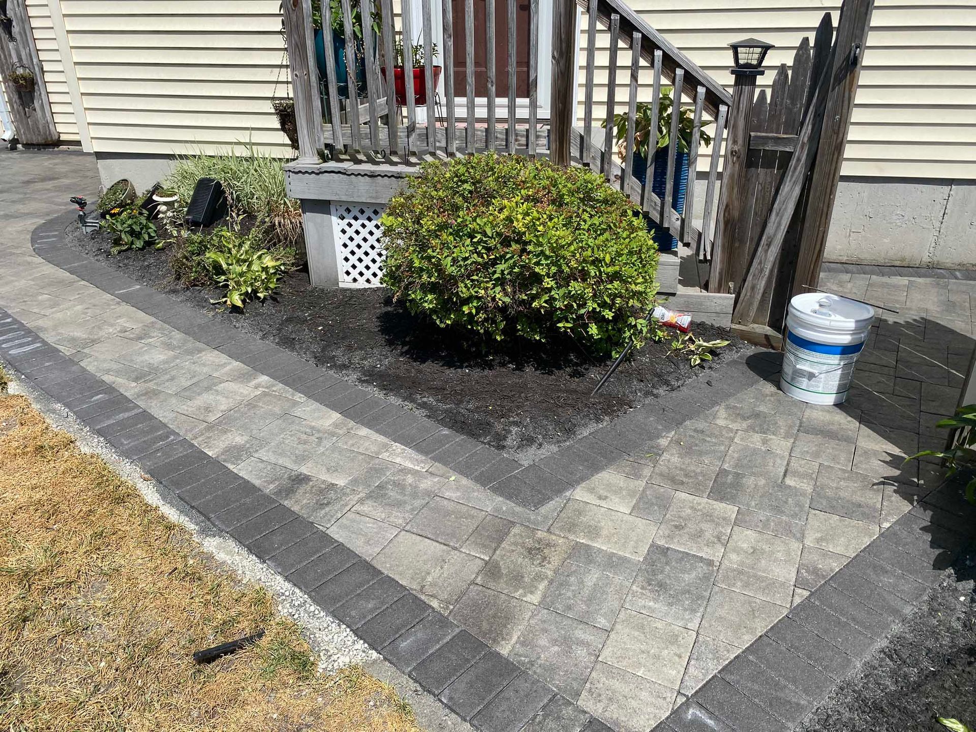 A patio made of light grey pavers with a dark charcoal border, fronting a house entrance with steps and a small garden.