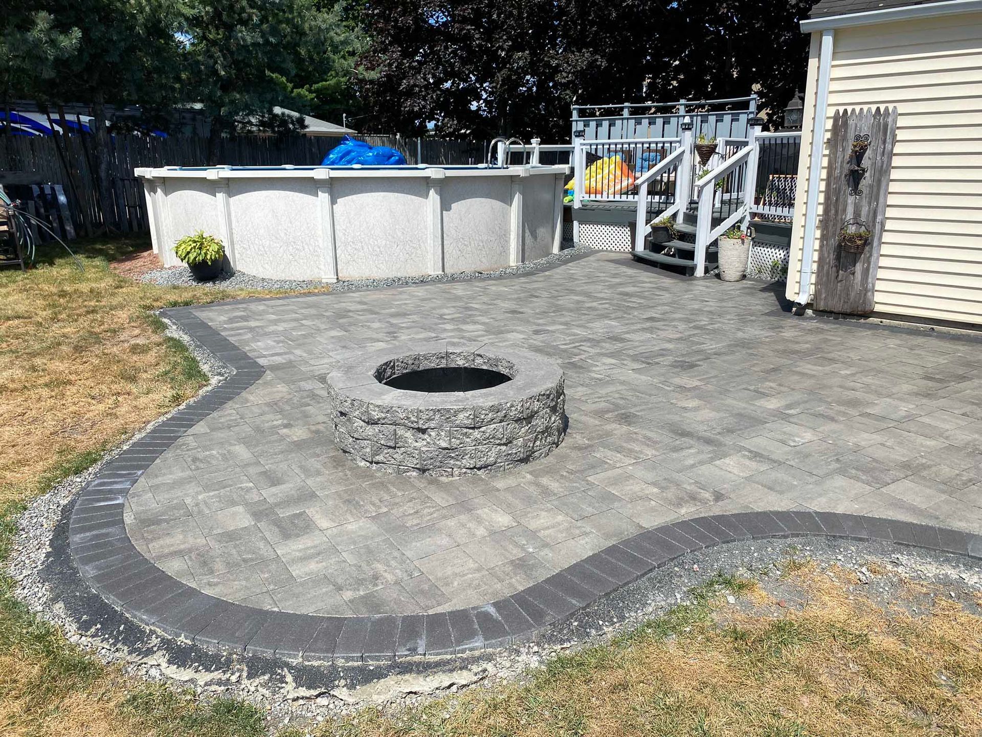 A stone fire pit centered on a gray paver patio next to an above-ground swimming pool in a backyard.