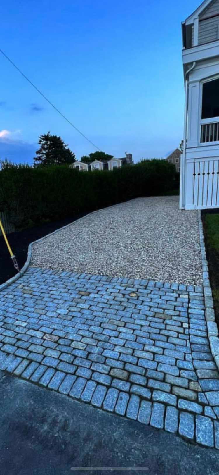 A stone paver apron leads into a gravel driveway next to a white building with a porch, framed by a dark green hedge.