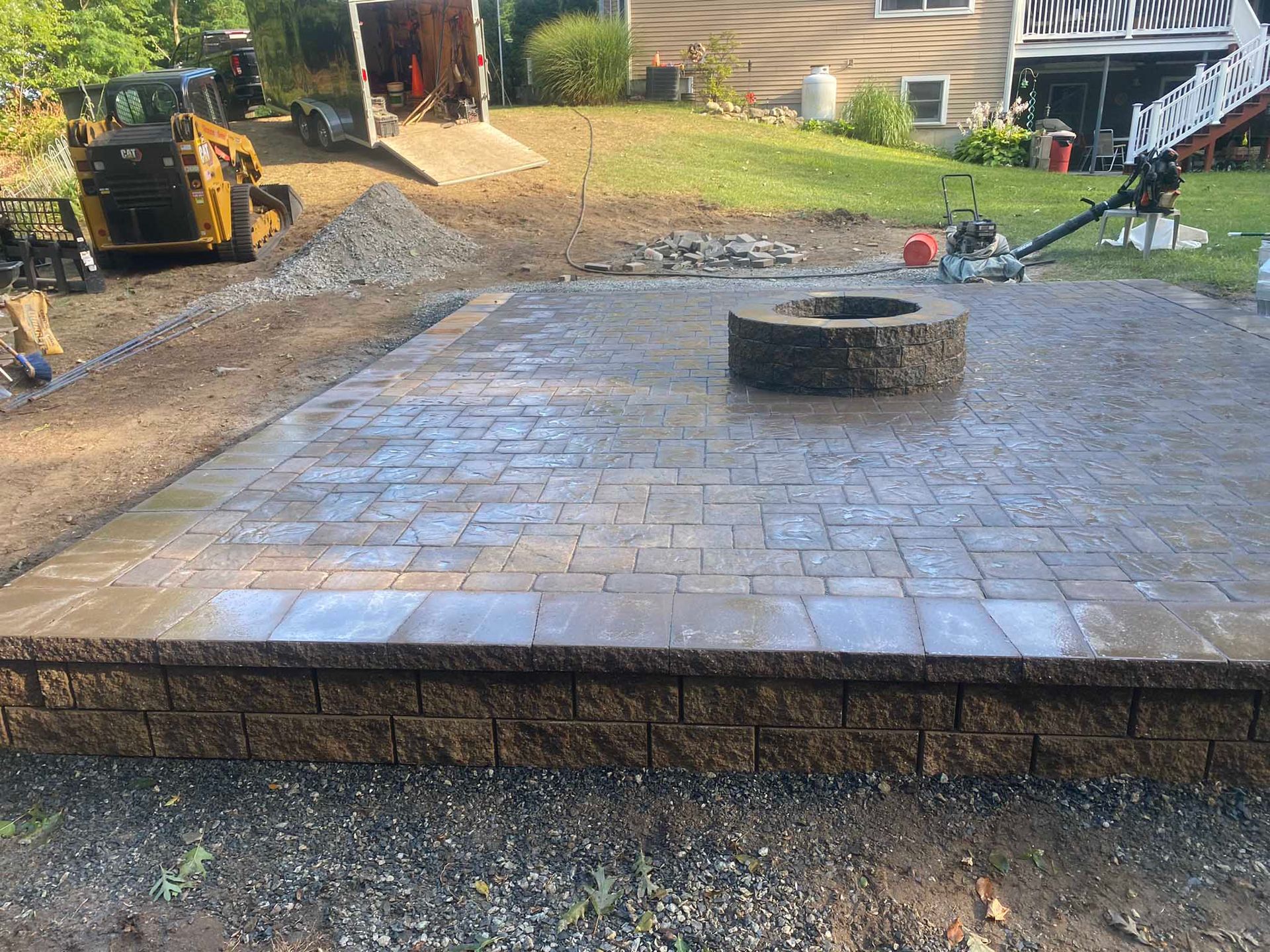 A newly constructed rectangular stone paver patio with a matching circular fire pit in a residential backyard.