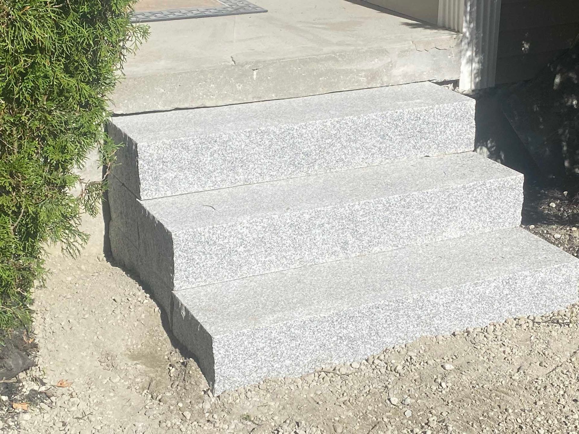 Three stone steps with a grey speckled texture leading up to a concrete porch next to a green bush.