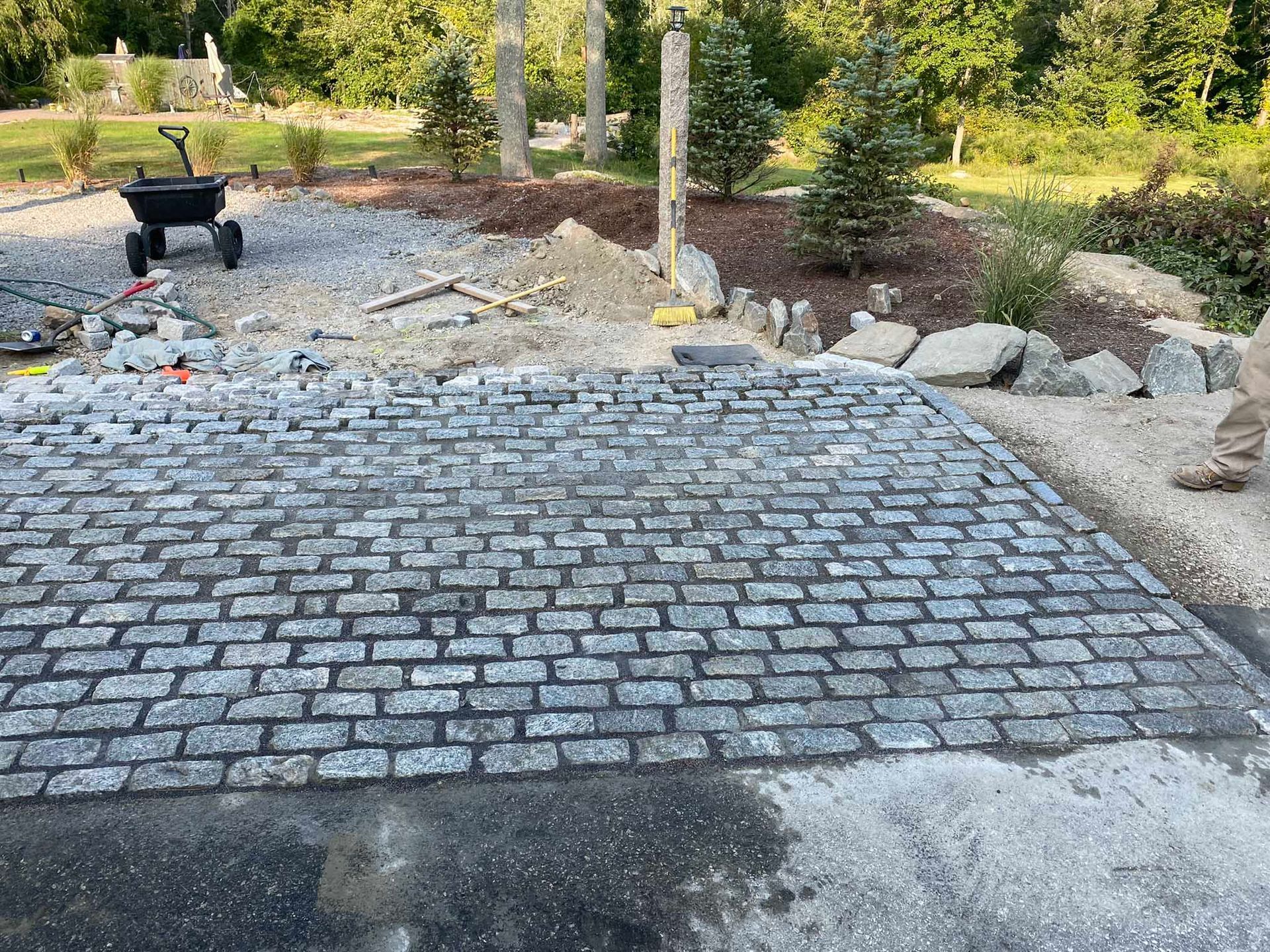 A rectangular area of newly laid grey granite cobblestones in a landscape setting with mulch and trees in the background.