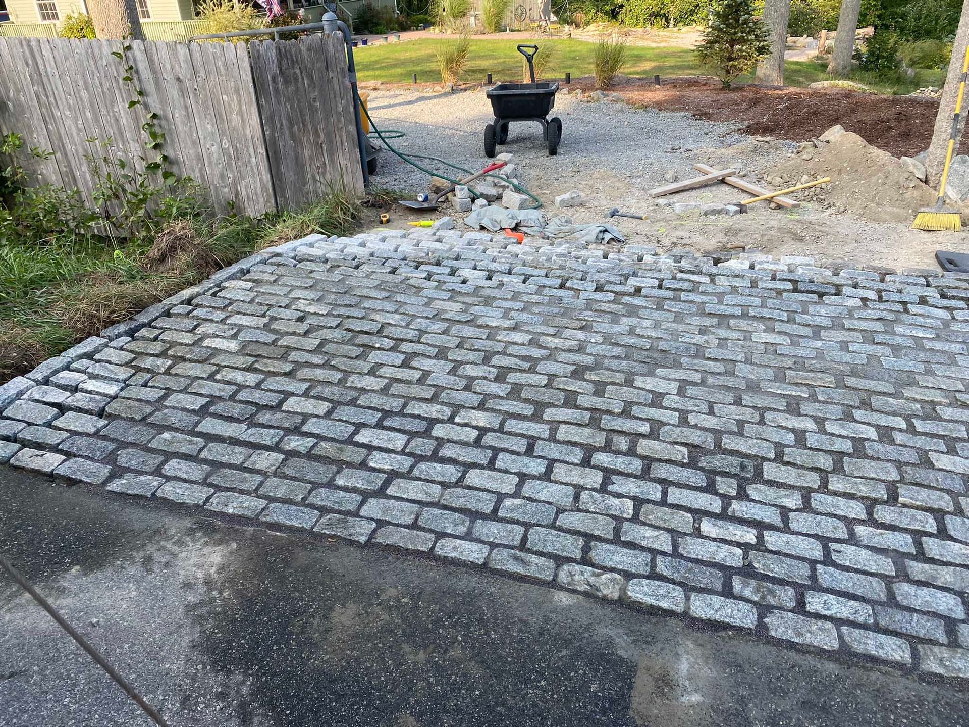 A newly installed gray cobblestone driveway apron transitions to a gravel yard with a small black utility cart.