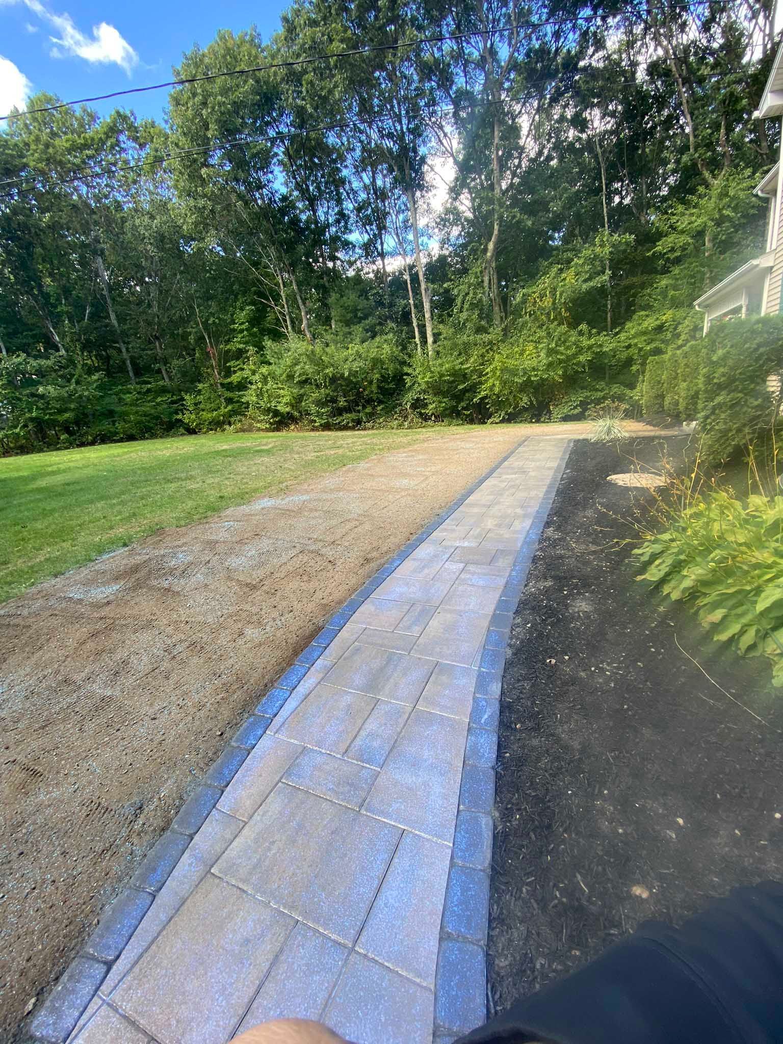A newly laid paver walkway with a blue border curves through a grassy yard alongside a dark mulched garden bed.