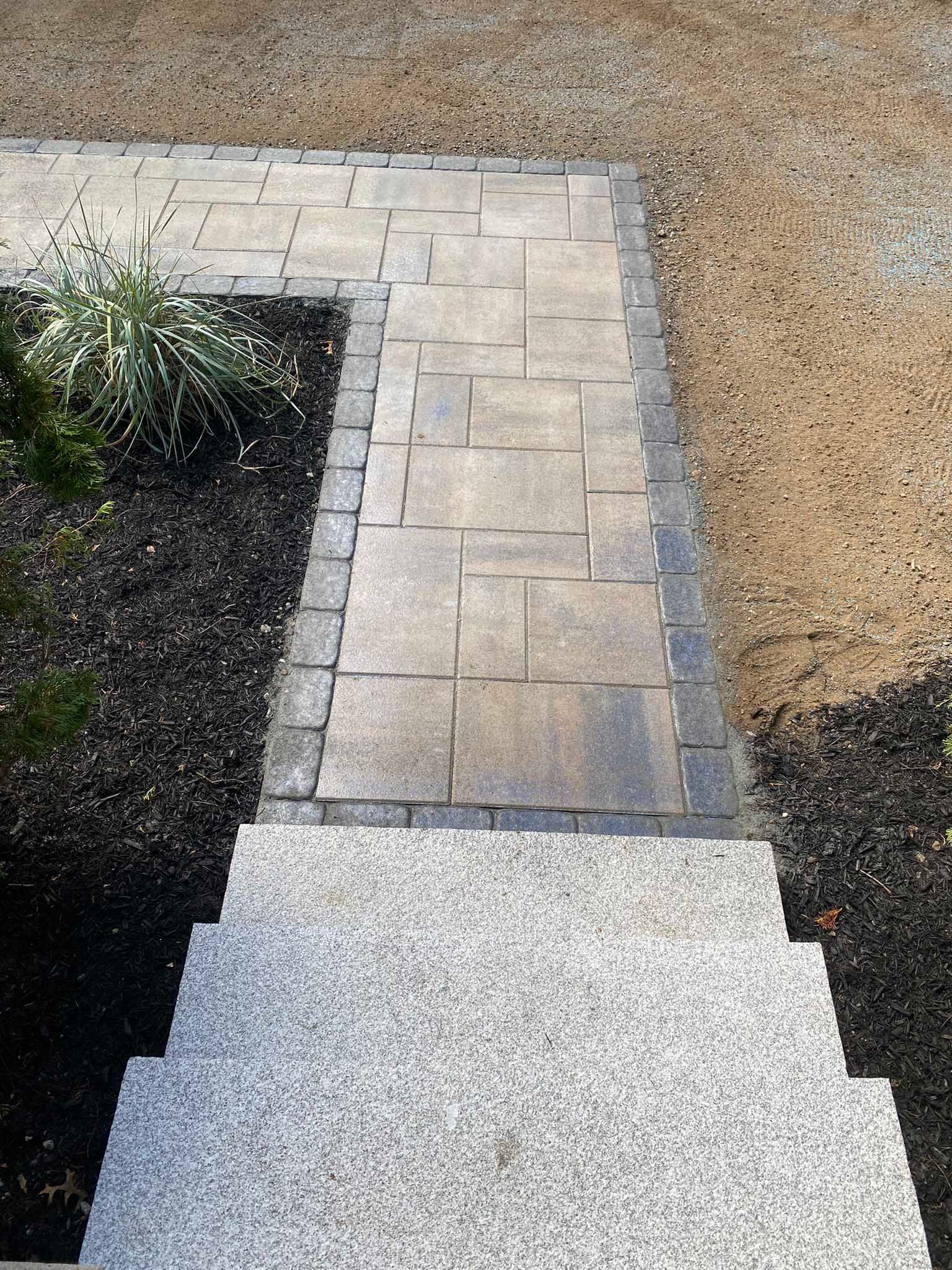 A top-down view of stone steps leading to a rectangular paver walkway bordered by dark bricks, set between mulched beds.