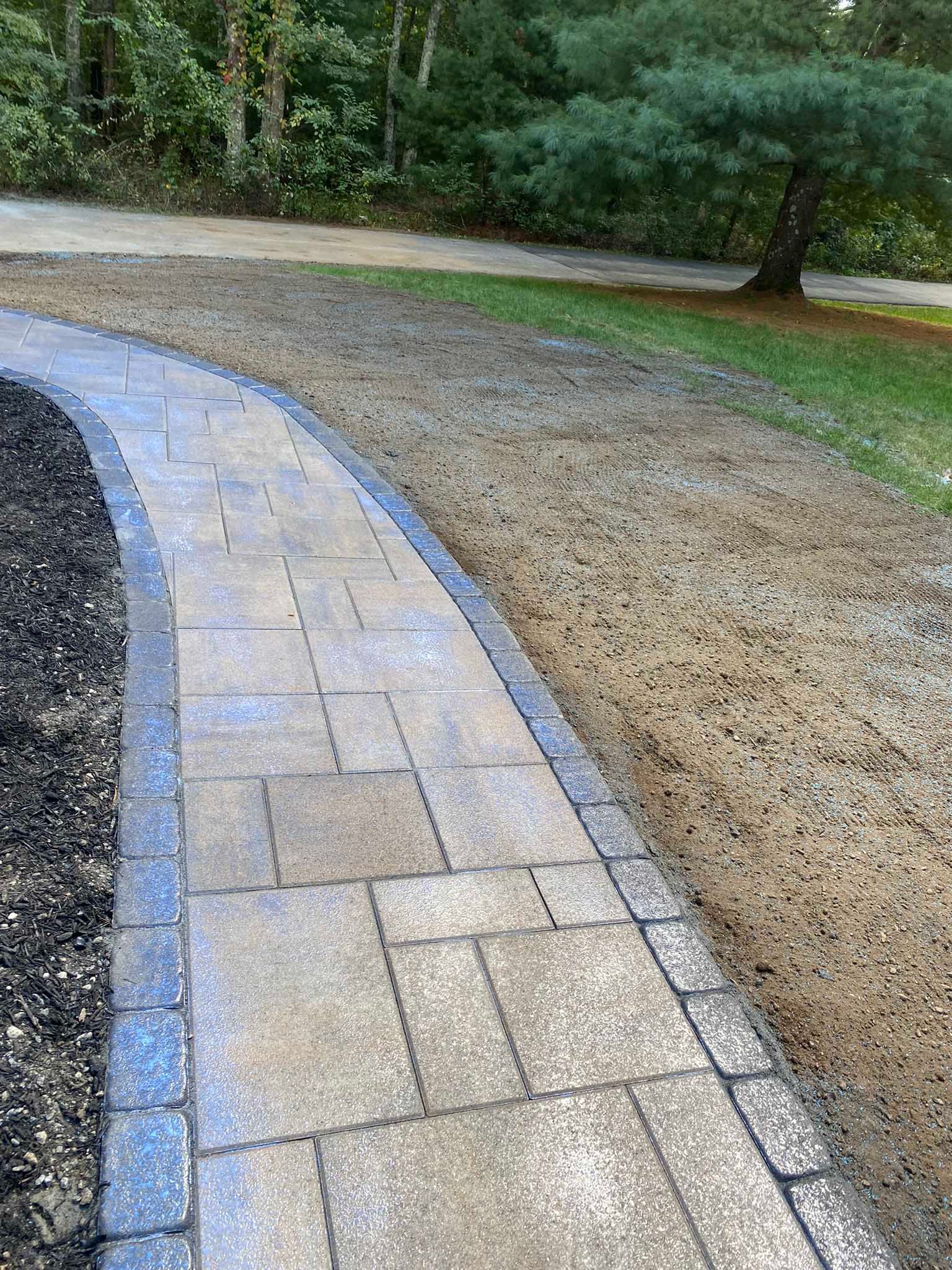 A curved paved walkway with a dark border runs between a dark gravel bed and a newly graded dirt area near trees.